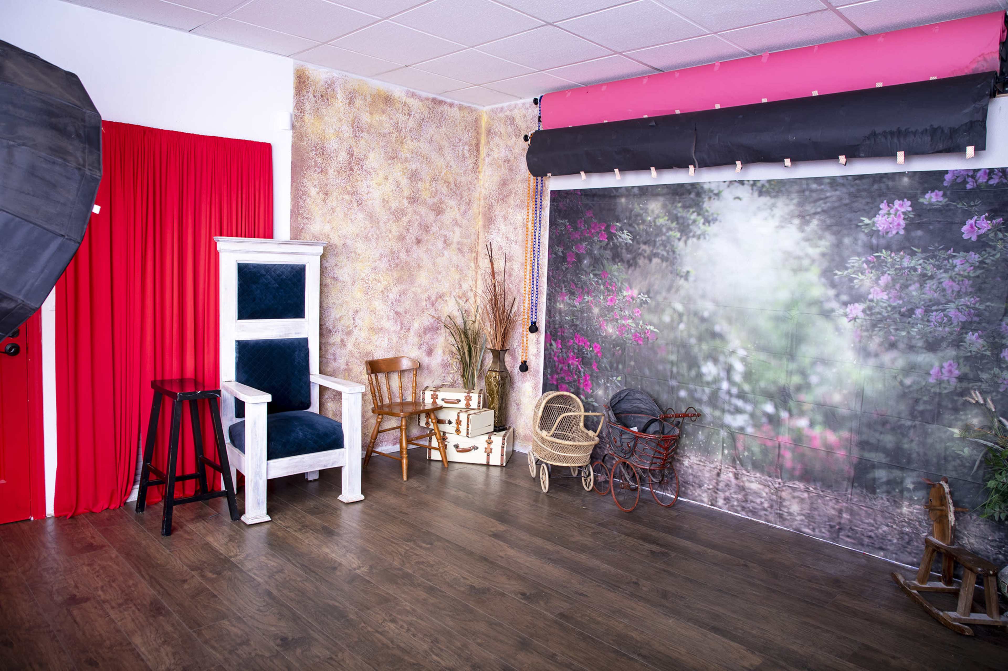 A photography studio featuring a blue velvet chair, a tall white throne-like chair, and various rustic props against a backdrop of floral scenery.