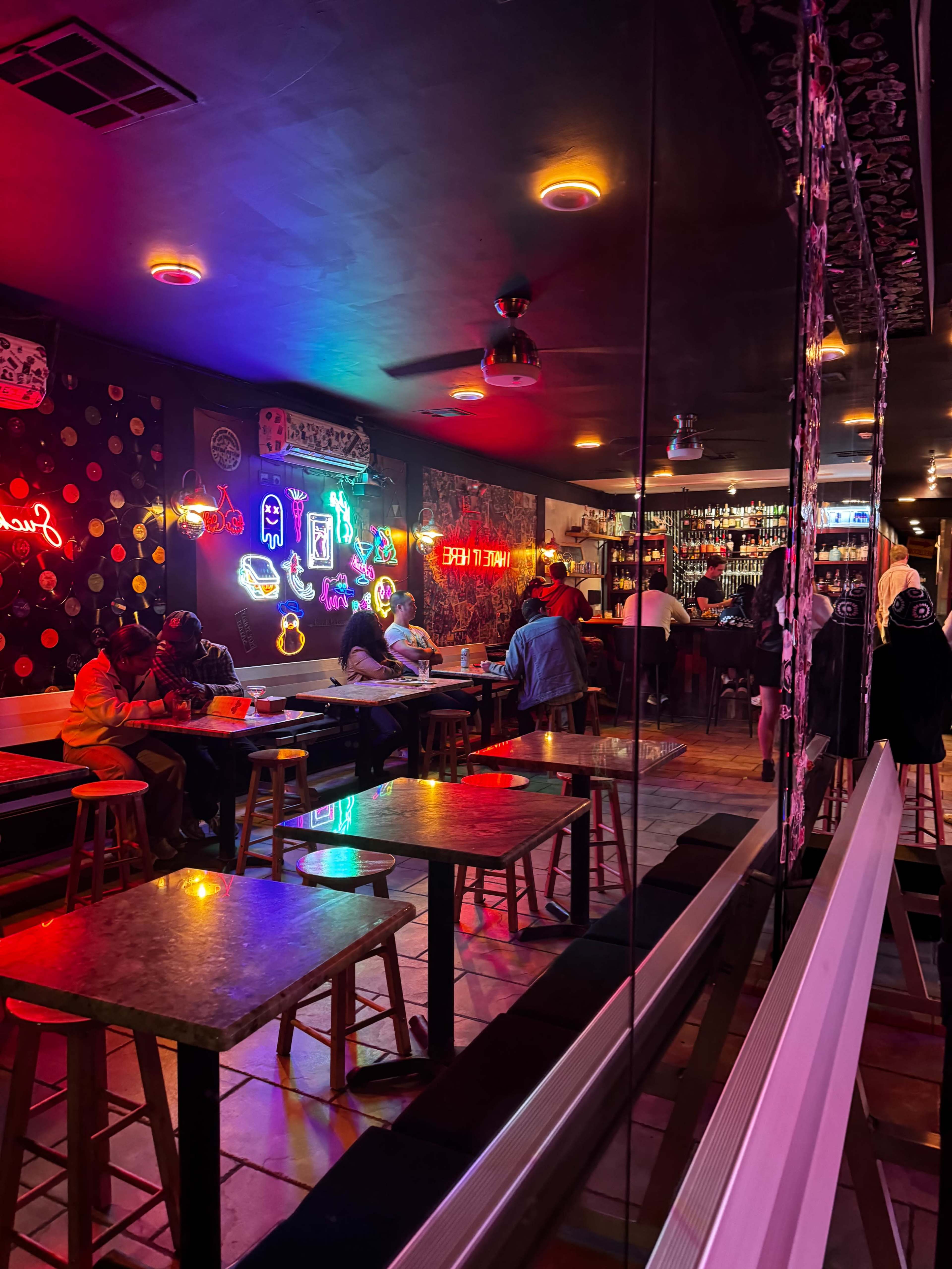 The image shows the interior of a vibrant bar with neon lights, wooden tables, and patrons seated at the bar and tables.