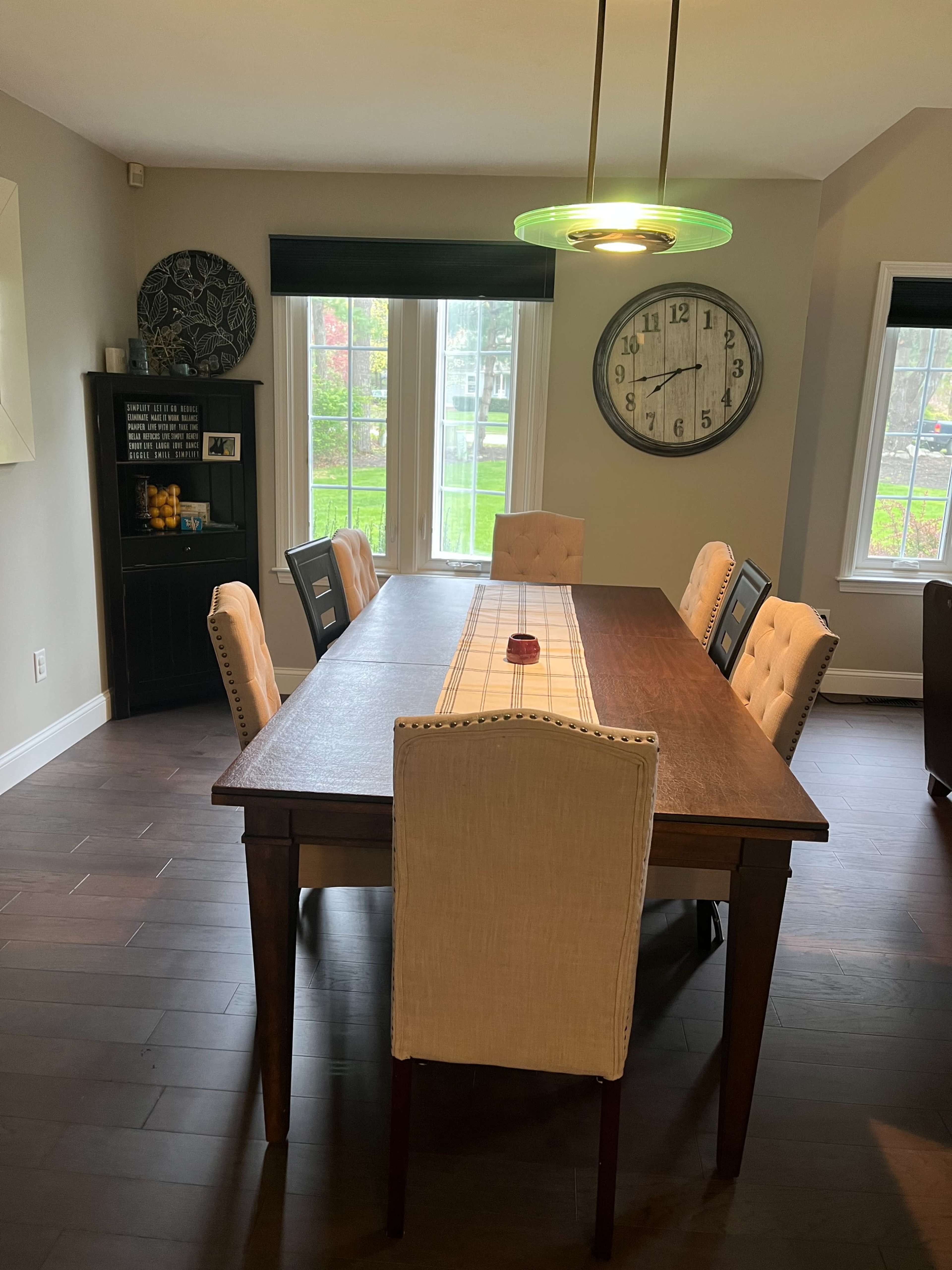 A modern dining room features a long wooden table surrounded by upholstered chairs, with a large clock and windows letting in natural light.