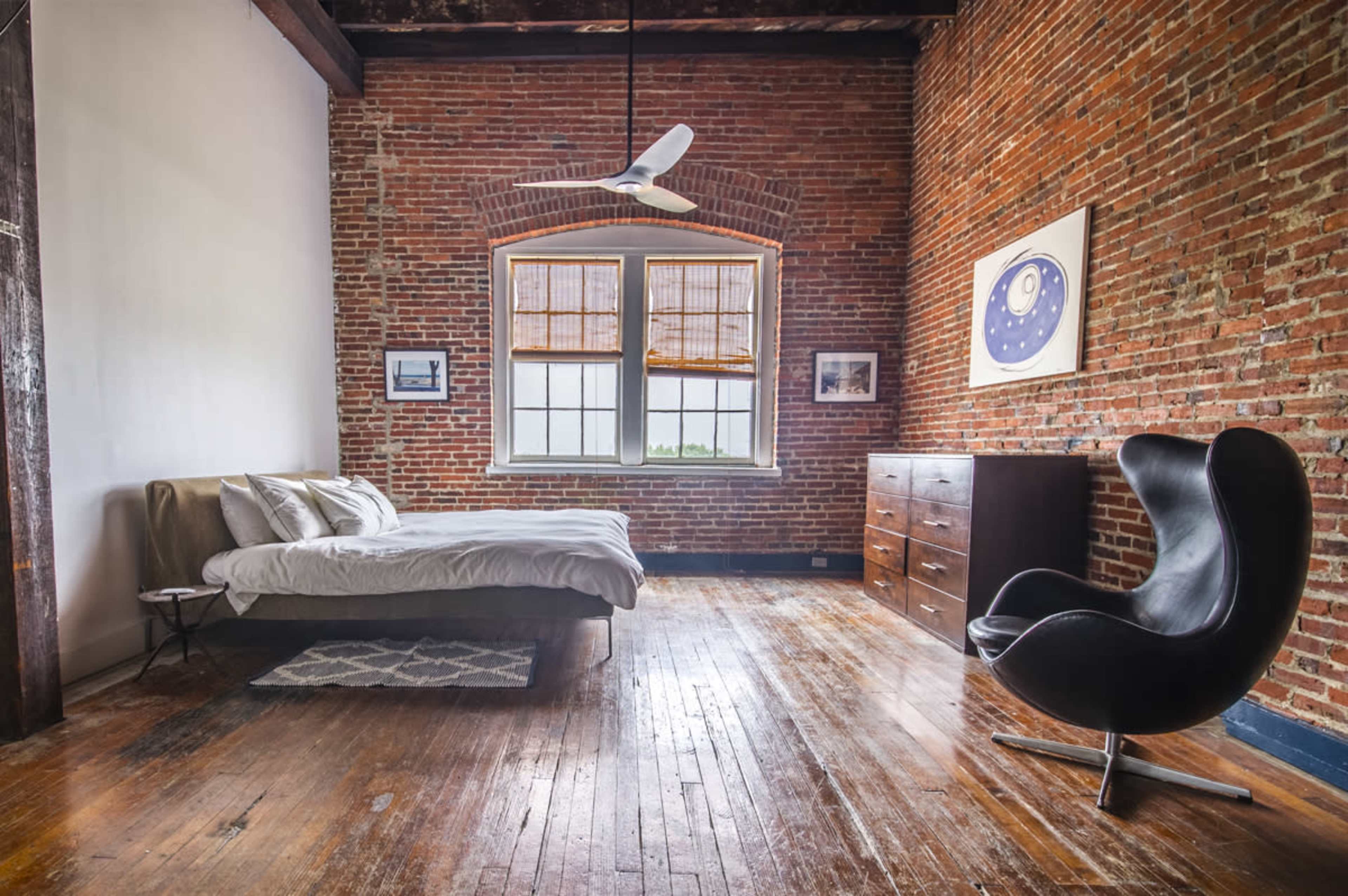 The image shows a minimalist bedroom with a bed, a wooden dresser, and a distinctive black chair, set against exposed brick walls and wooden flooring.