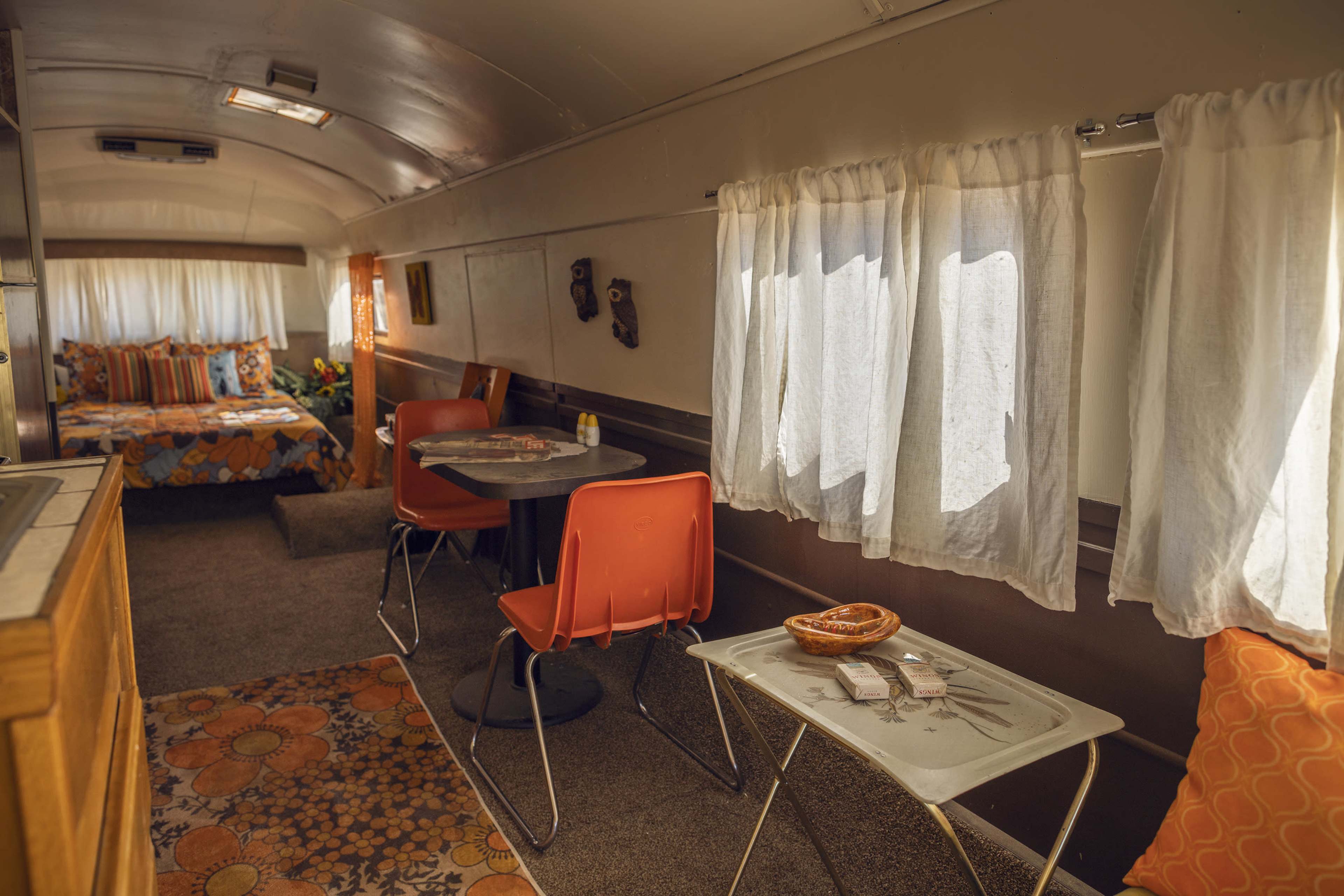 The interior of a vintage trailer featuring a dining area with orange chairs, a small table, and a bedroom area with a patterned bedspread.