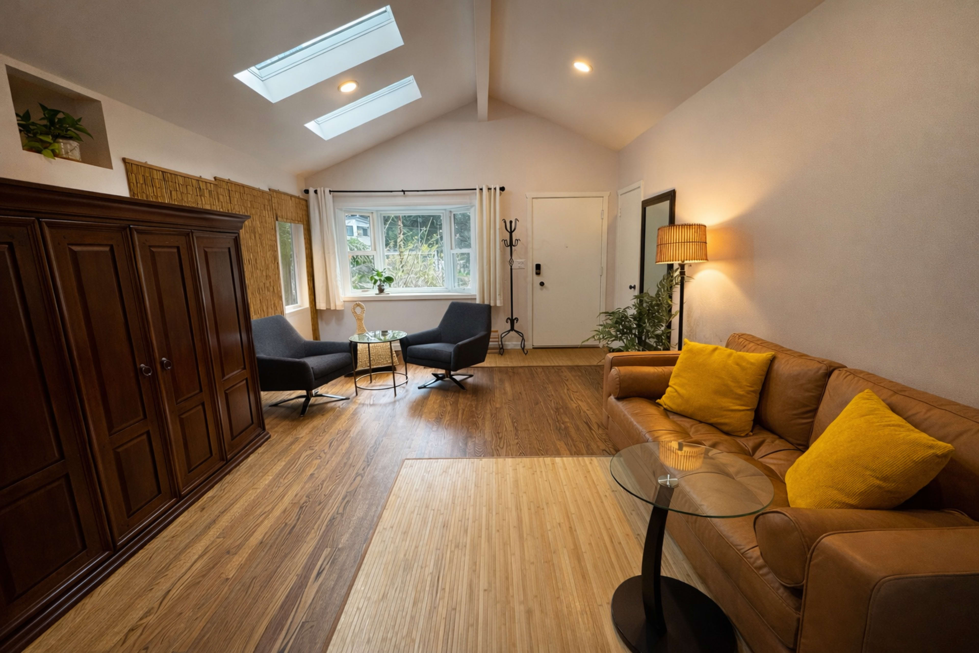 The image shows a cozy living area with a brown leather couch, two gray armchairs, a small round table, and large windows allowing natural light in.