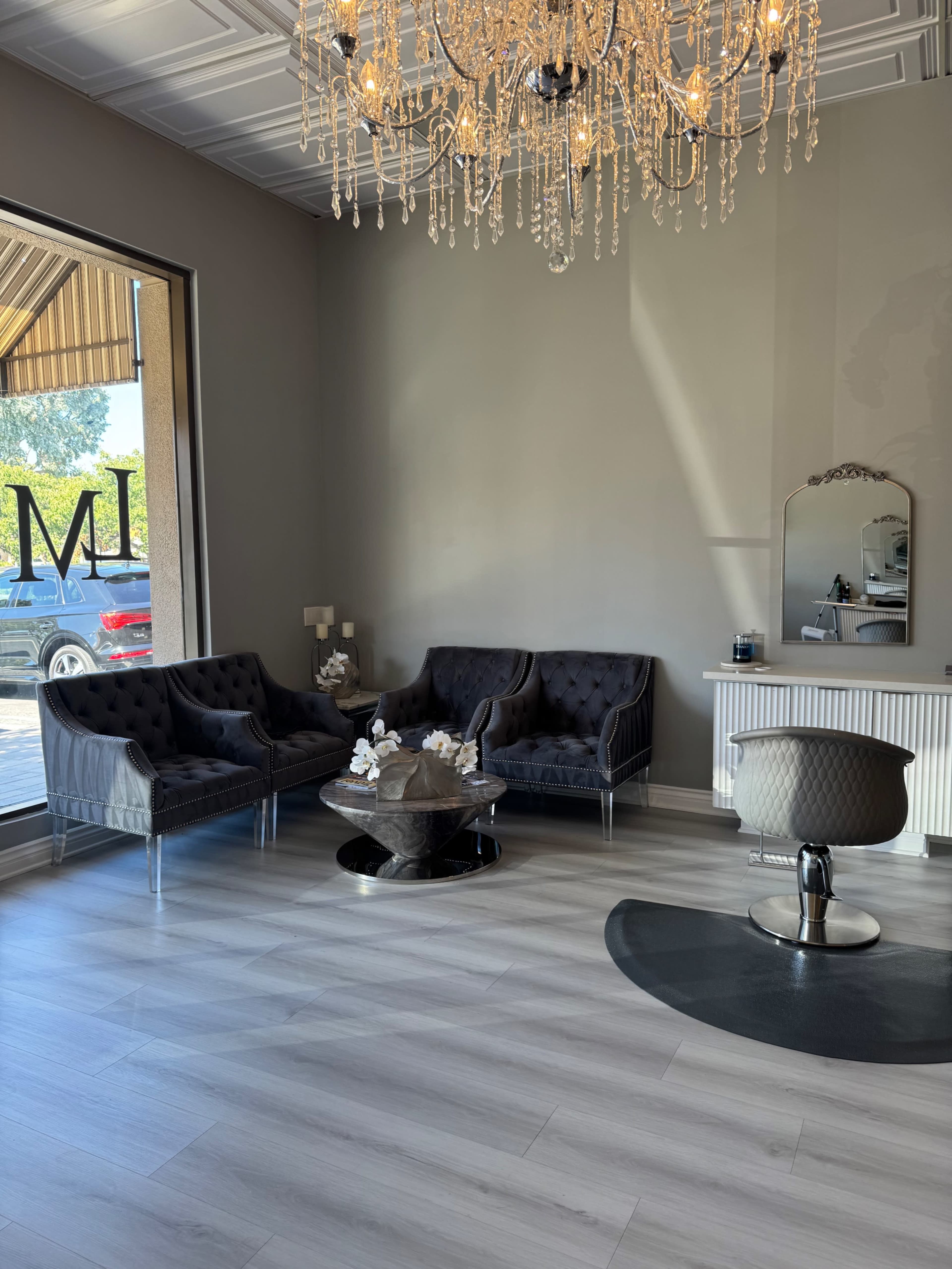 The image shows a modern salon interior with plush seating, a round glass coffee table, a mirror, and a large chandelier overhead.