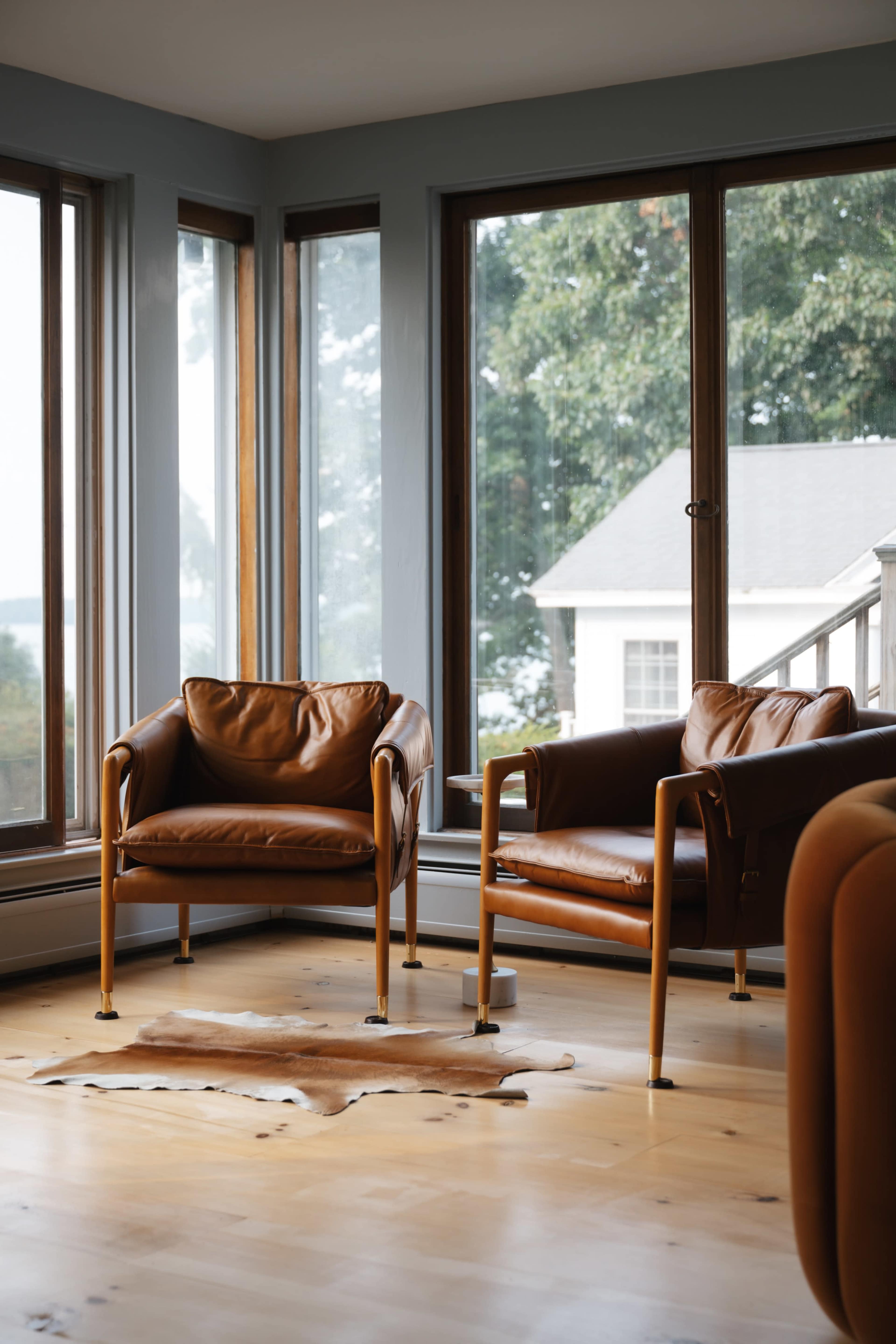 Two brown leather chairs are positioned in a bright room with large windows providing views of the outdoors.