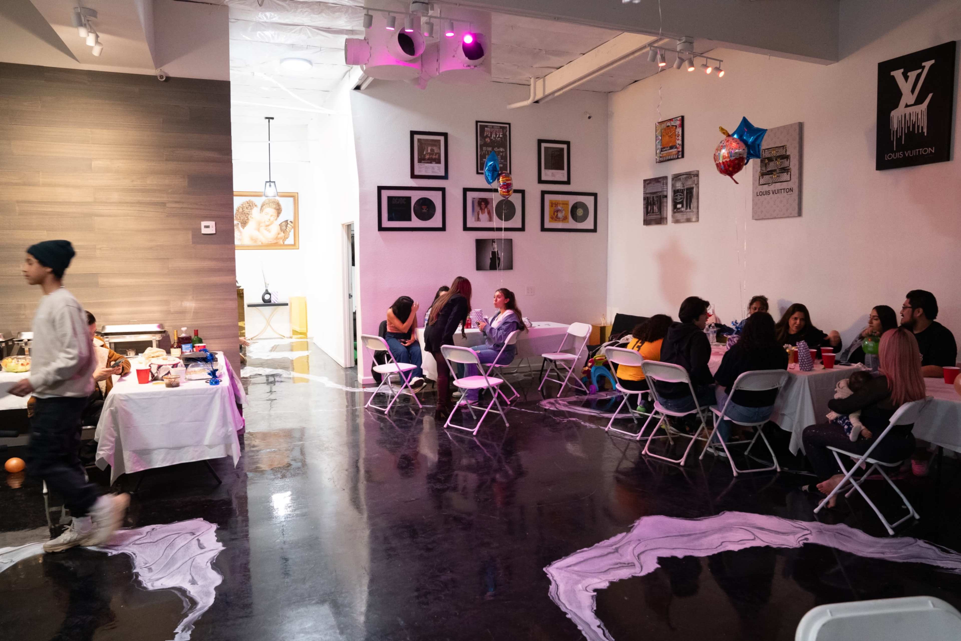 A spacious indoor event venue filled with people seated at tables and a buffet, decorated with balloons and artwork on the walls.