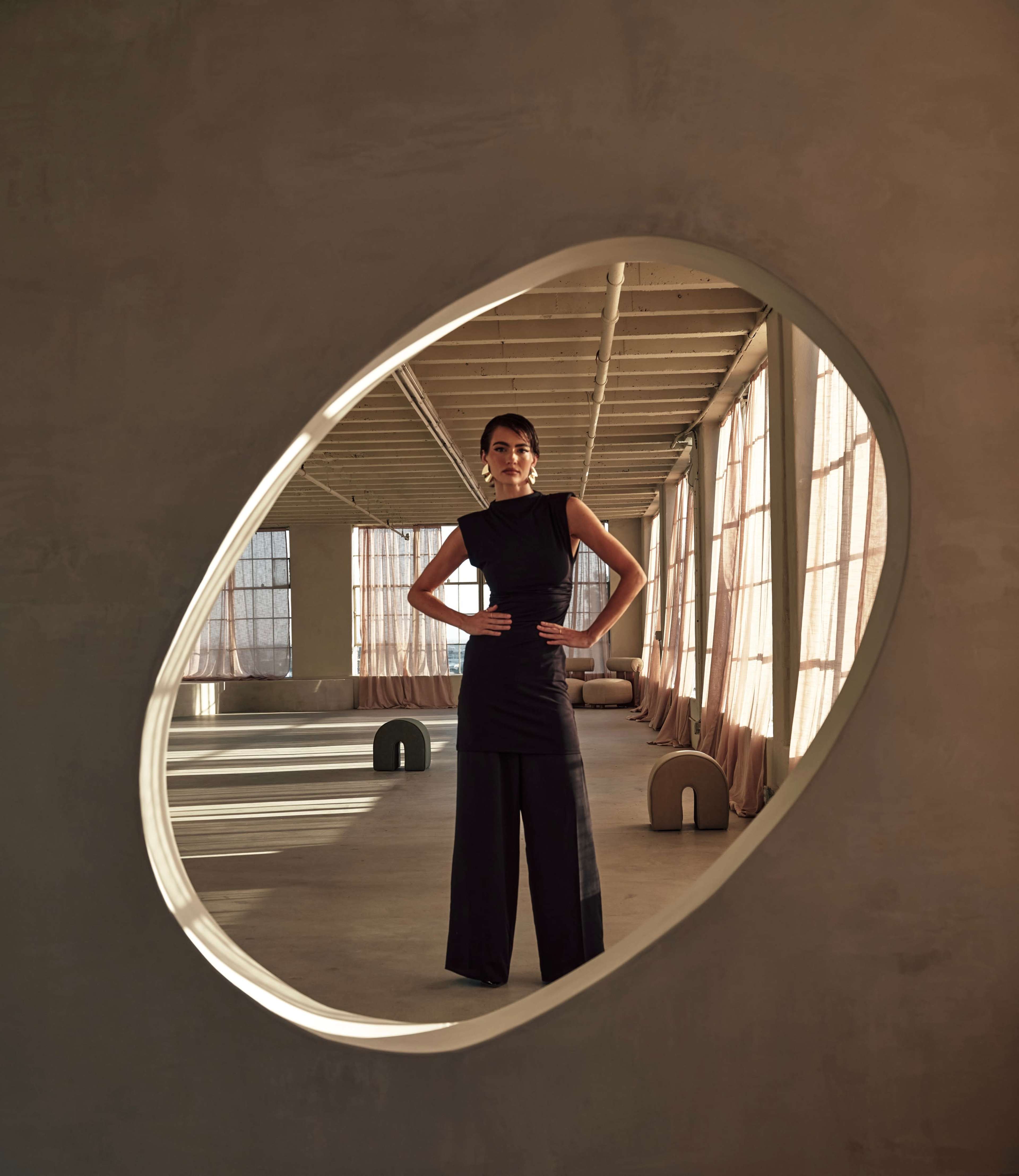 A woman poses confidently in a black outfit inside a spacious, minimalist room framed by an oval cutout opening.