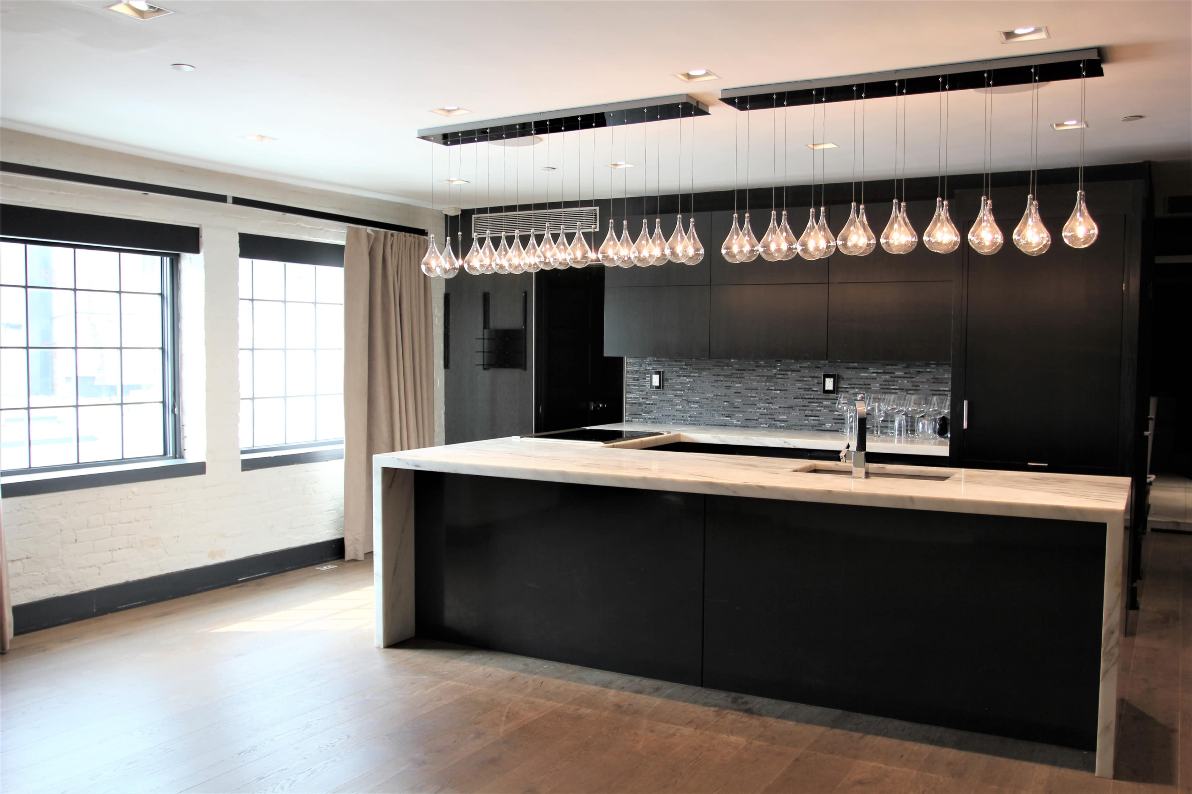 A modern kitchen featuring a large island with a marble countertop, dark cabinetry, and a row of glass pendant lights hanging overhead.