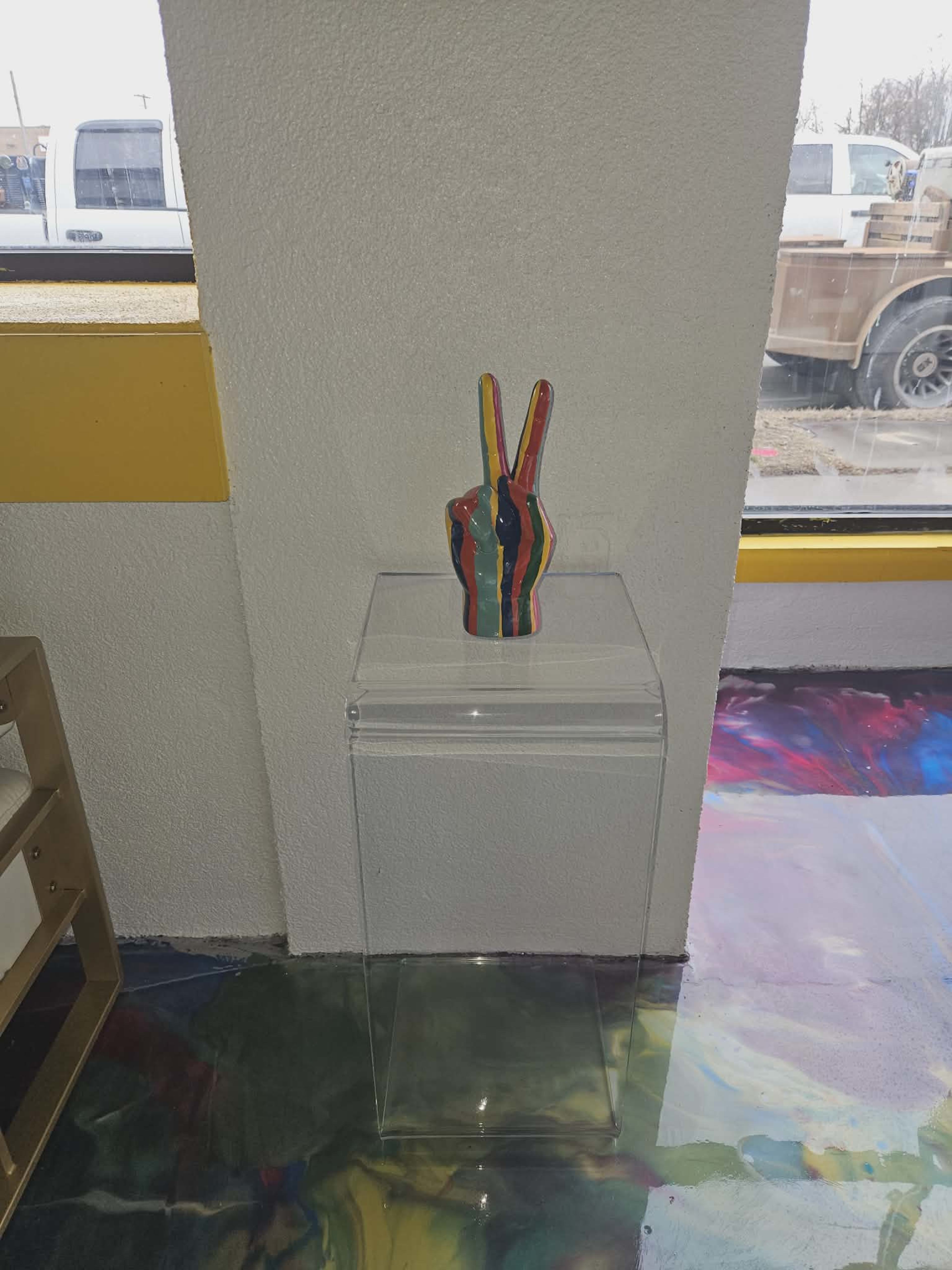 A colorful peace sign sculpture is displayed on a clear pedestal in a brightly lit room with a multicolored floor.