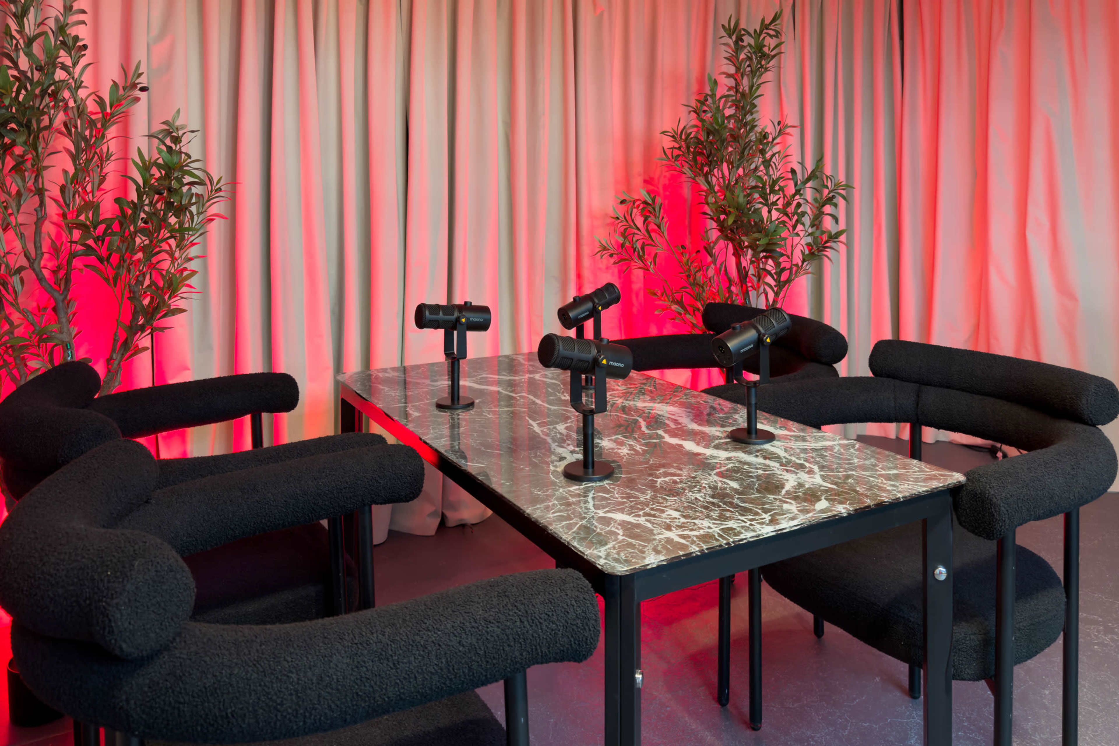 A modern conference table with a marble top is surrounded by black, curved seating and equipped with four microphones, set against a backdrop of red-lit curtains and potted plants.
