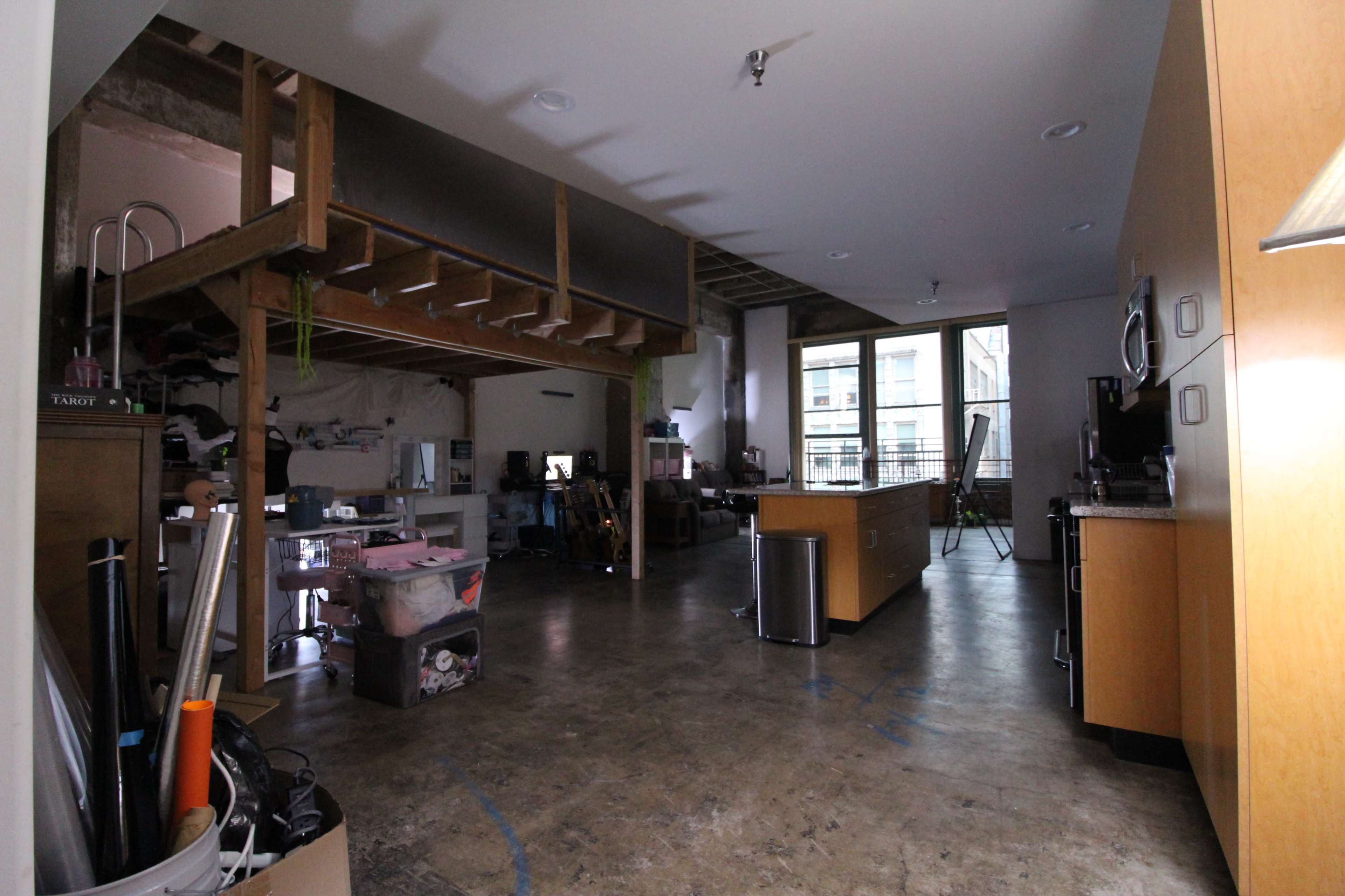 The image shows an open, industrial-style living space with a lofted area, kitchen island, and various items scattered throughout.