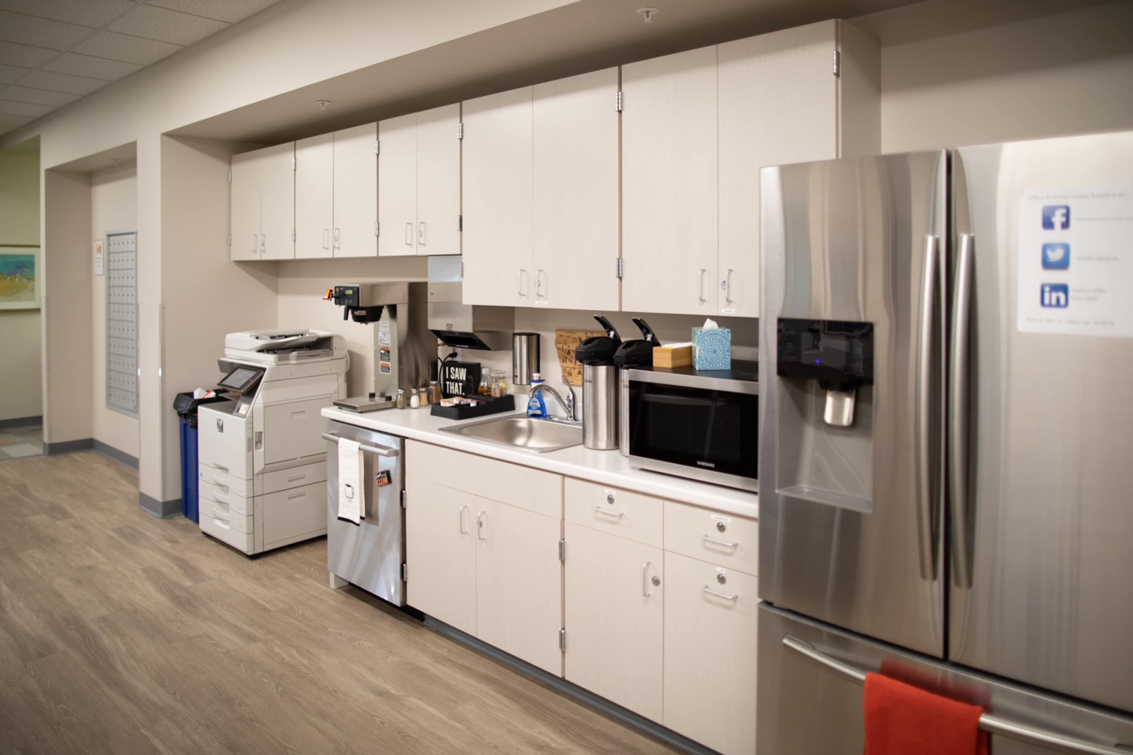 A modern office kitchen area with cabinets, a sink, a coffee machine, a microwave, and a stainless steel refrigerator.