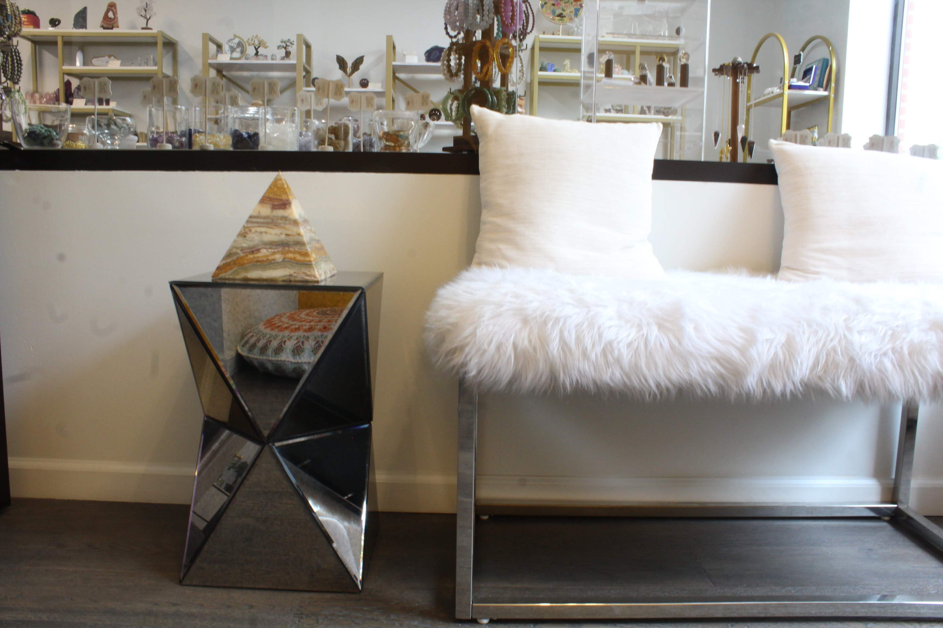 A mirrored geometric side table with a pyramidal decoration sits next to a white, fluffy bench adorned with pillows.
