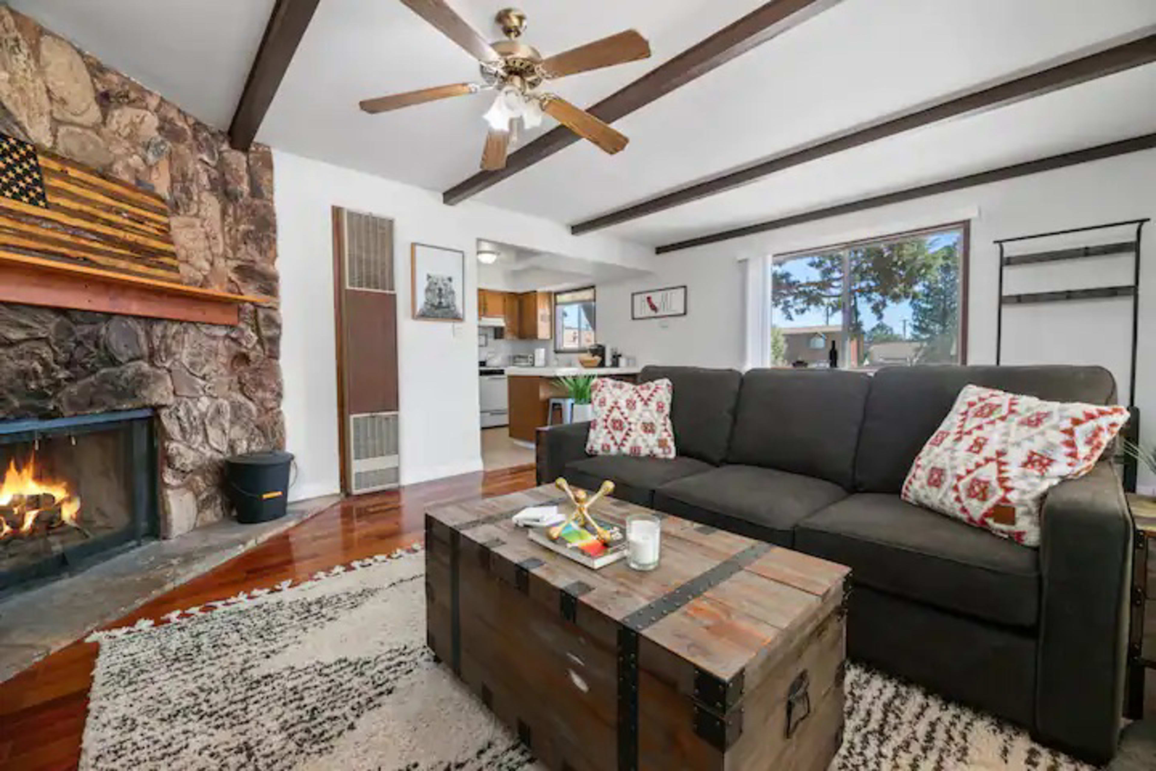 A cozy living room features a dark sofa, a wooden coffee table, and a stone fireplace, with an American flag art piece on the wall and a view of the outdoors through a window.