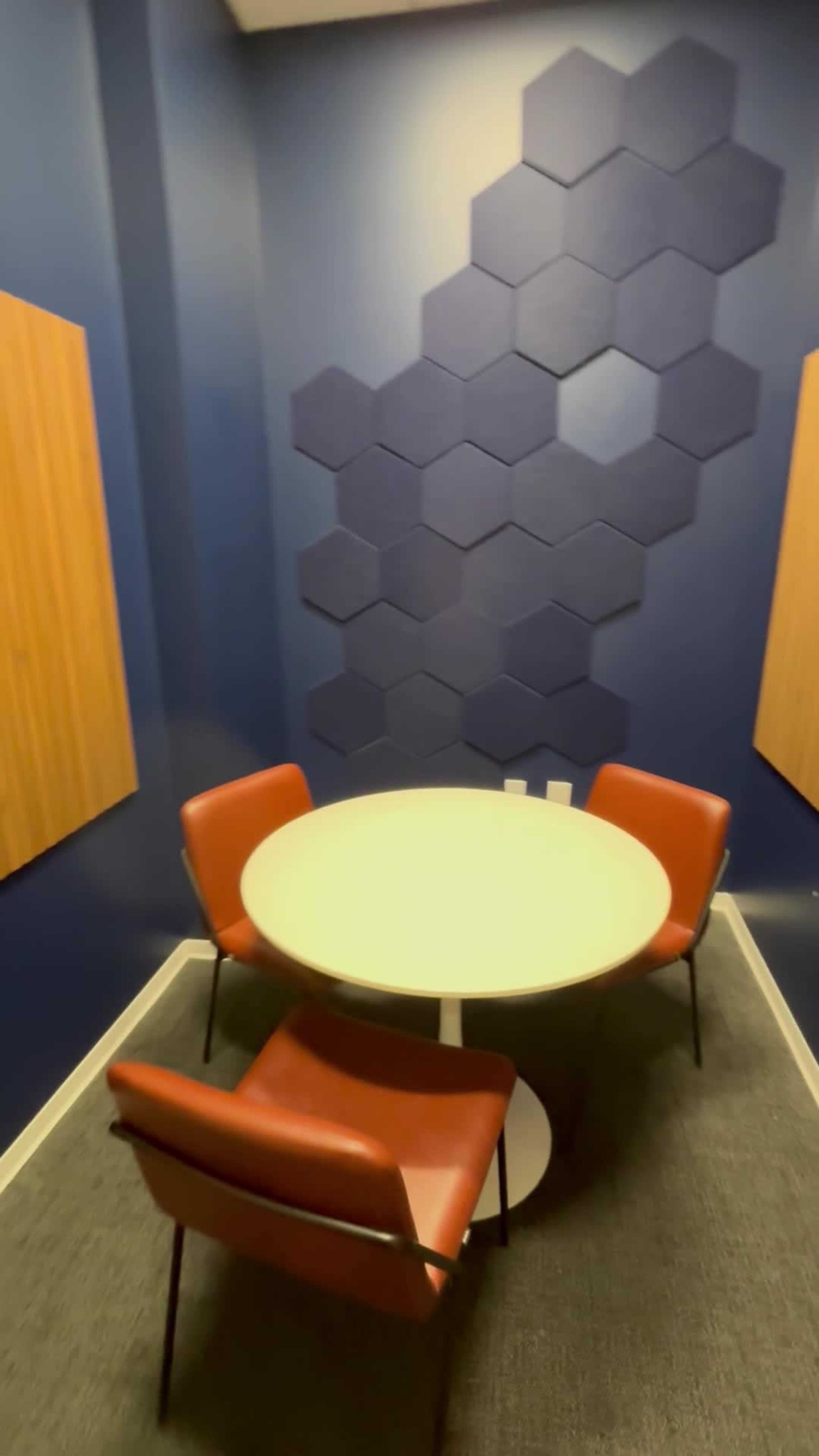 The image shows a small meeting room with a round table surrounded by four orange chairs and a wall featuring a hexagonal pattern.