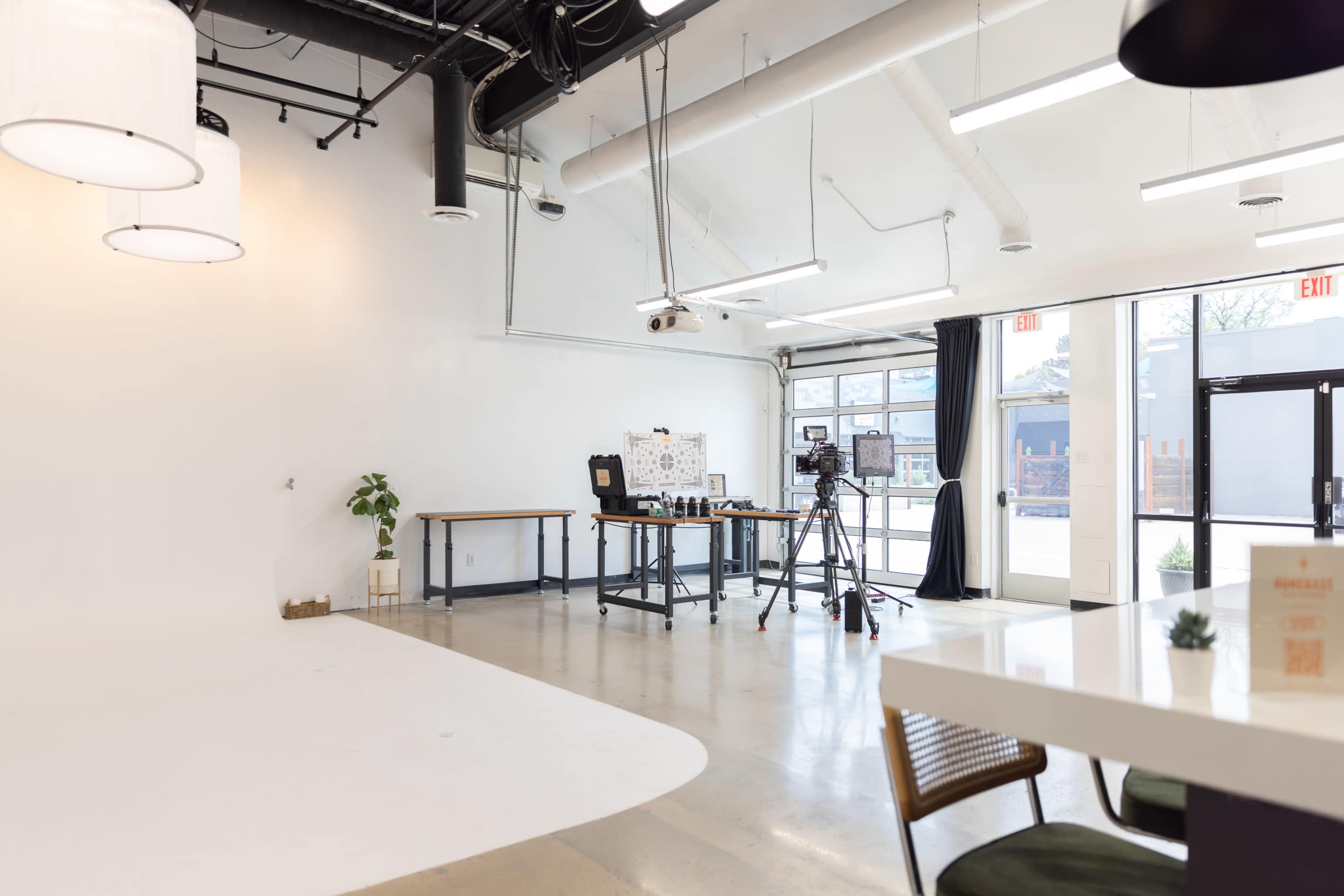 The image shows a spacious studio with a white backdrop, a table set up with photography equipment, and large windows allowing natural light to enter.