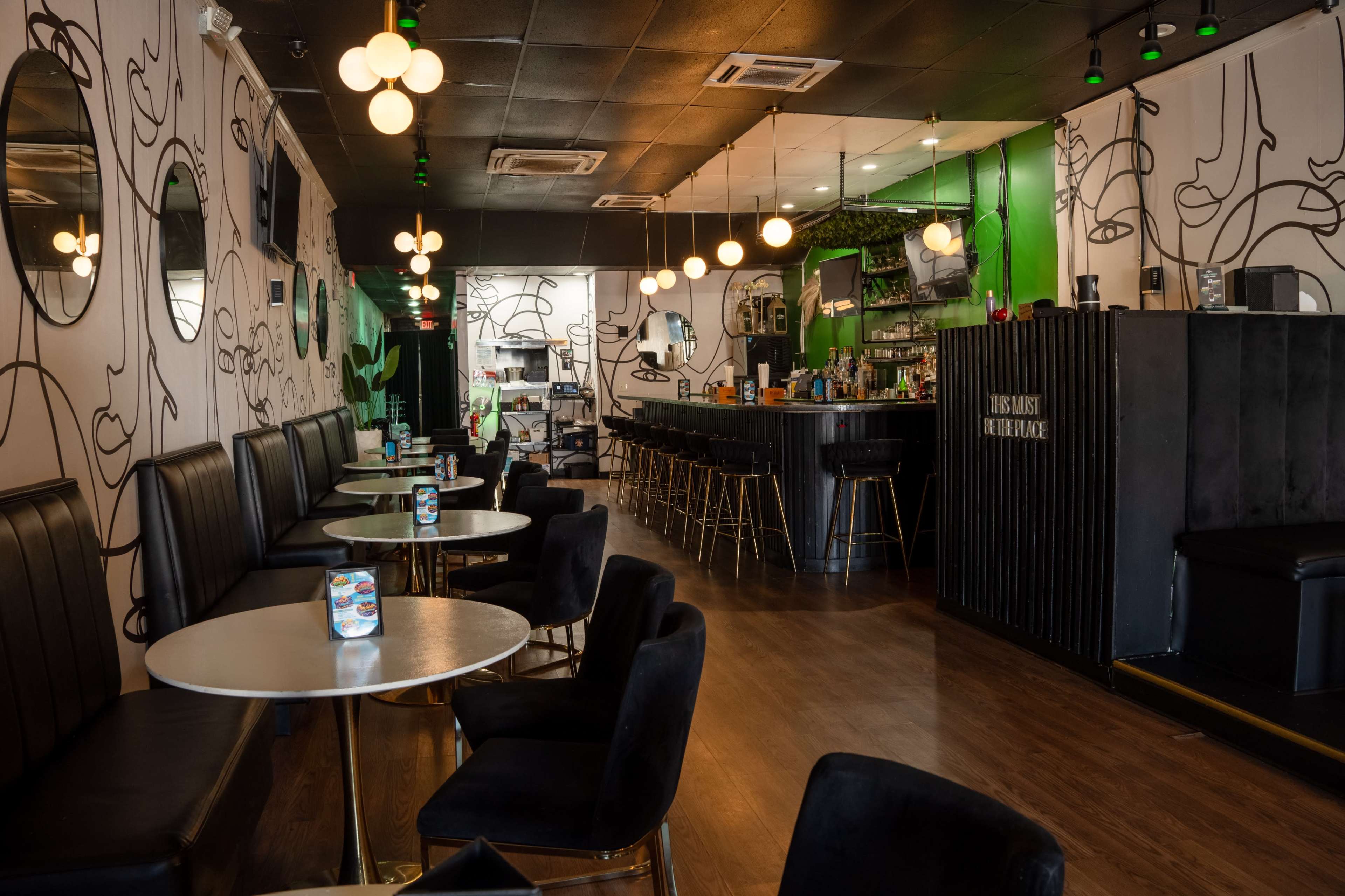 The image shows a modern bar or café interior with black seating, round tables, and decorative wall art, featuring a well-stocked bar in the background.