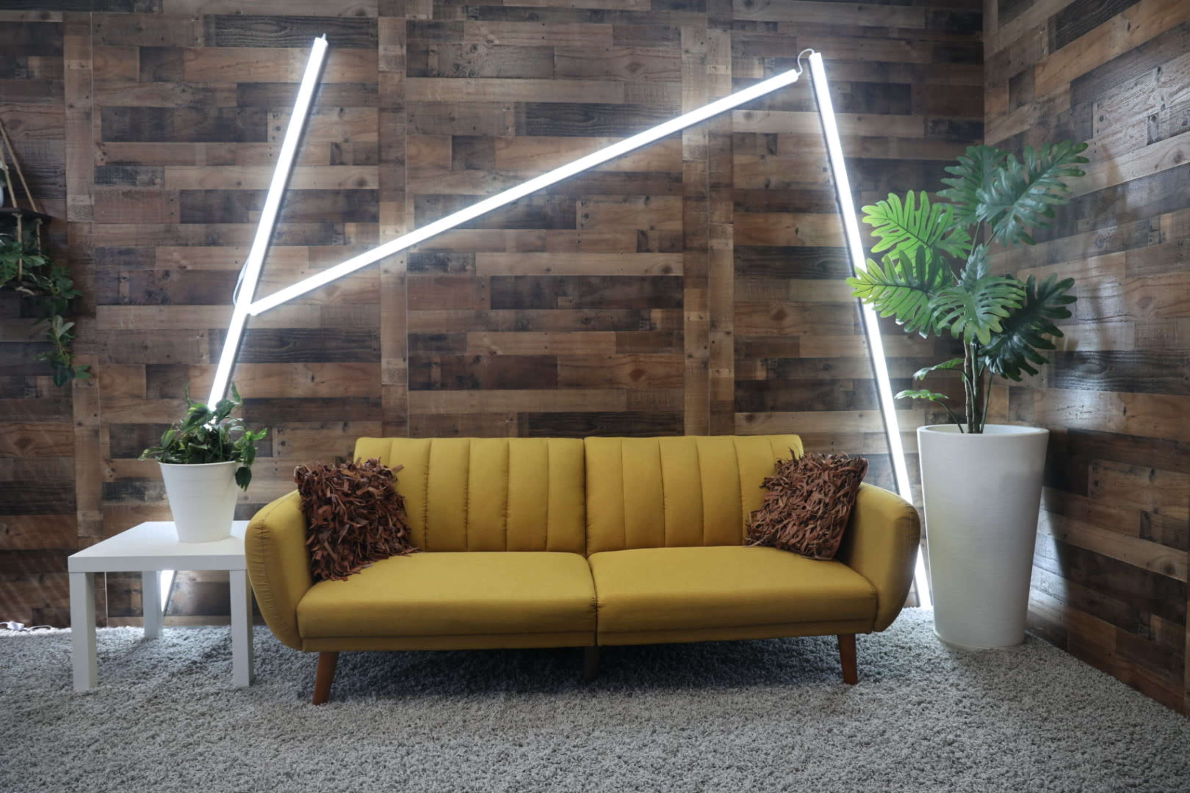 A mustard-colored sofa with decorative pillows is positioned in front of a wooden wall, accompanied by a tall plant and a small white table featuring a potted plant, with bright LED light strips creating a geometric pattern above.