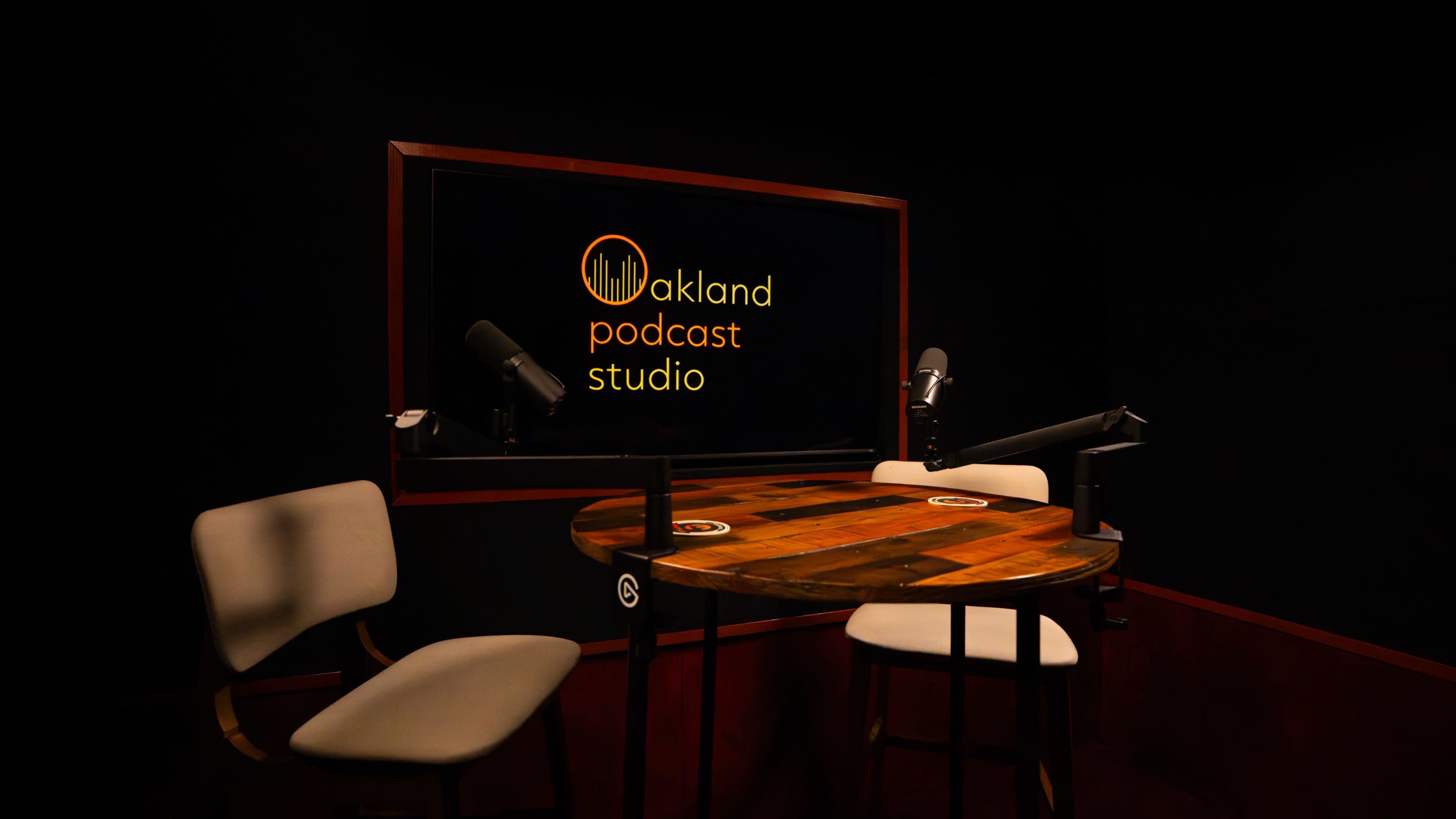 The image shows a dimly lit podcast studio with a wooden table and two high chairs, featuring microphones and a screen displaying the logo "Oakland Podcast Studio."
