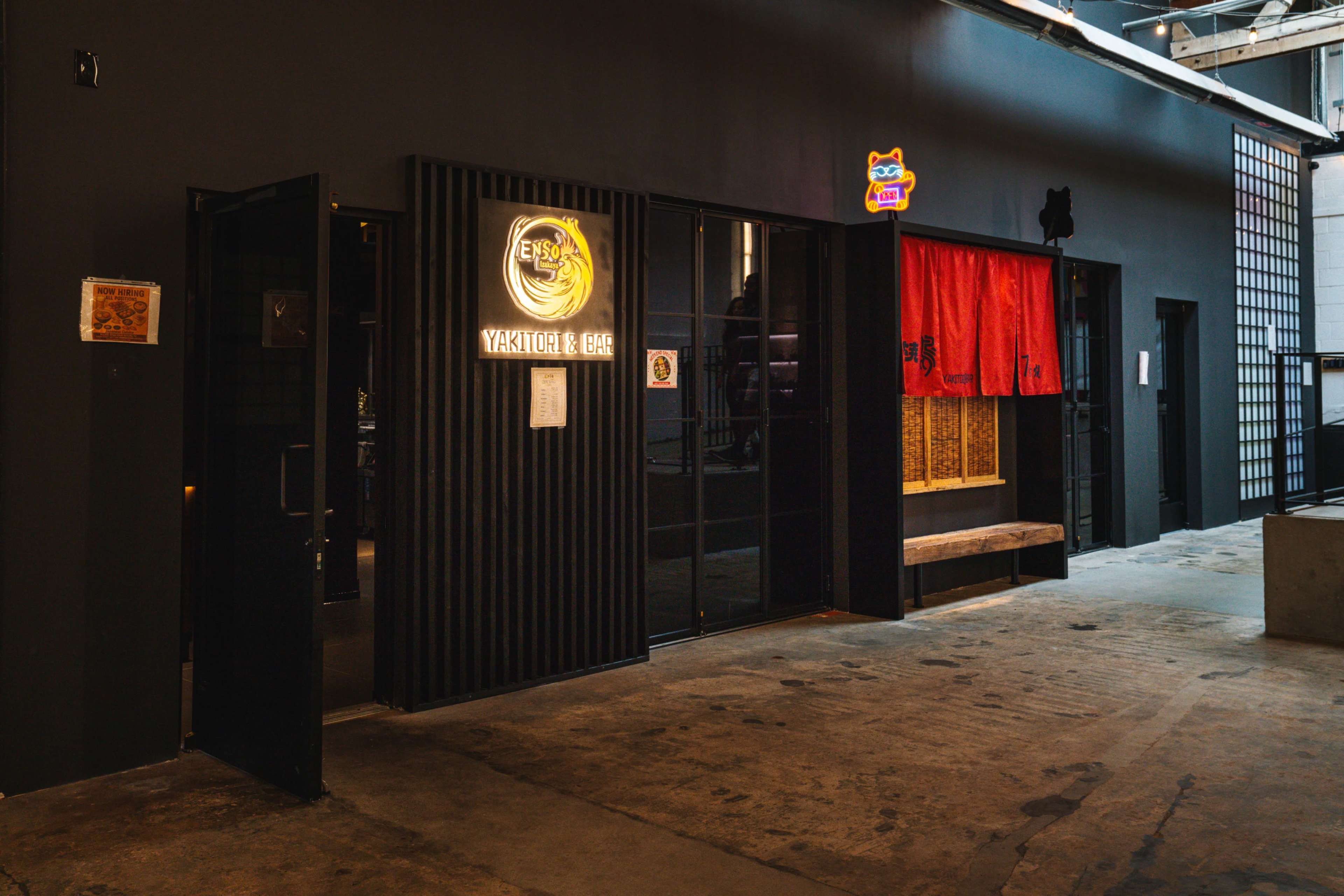 The image shows the entrance of a restaurant with a black exterior, featuring a lit sign that says "Yakitori & Bar," and a red fabric curtain partially covering the doorway.