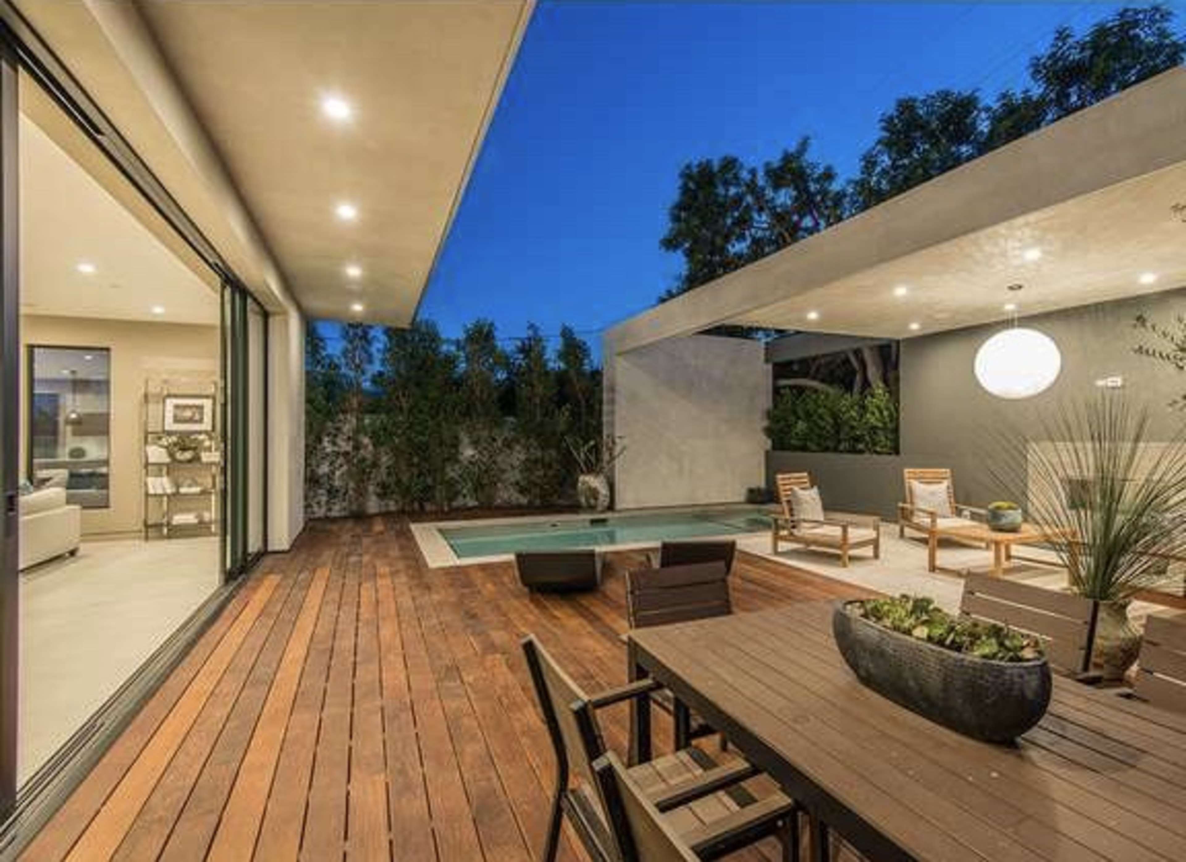 The image shows a modern outdoor patio with a wooden deck, a swimming pool, and sleek furniture under a softly lit overhead fixture.