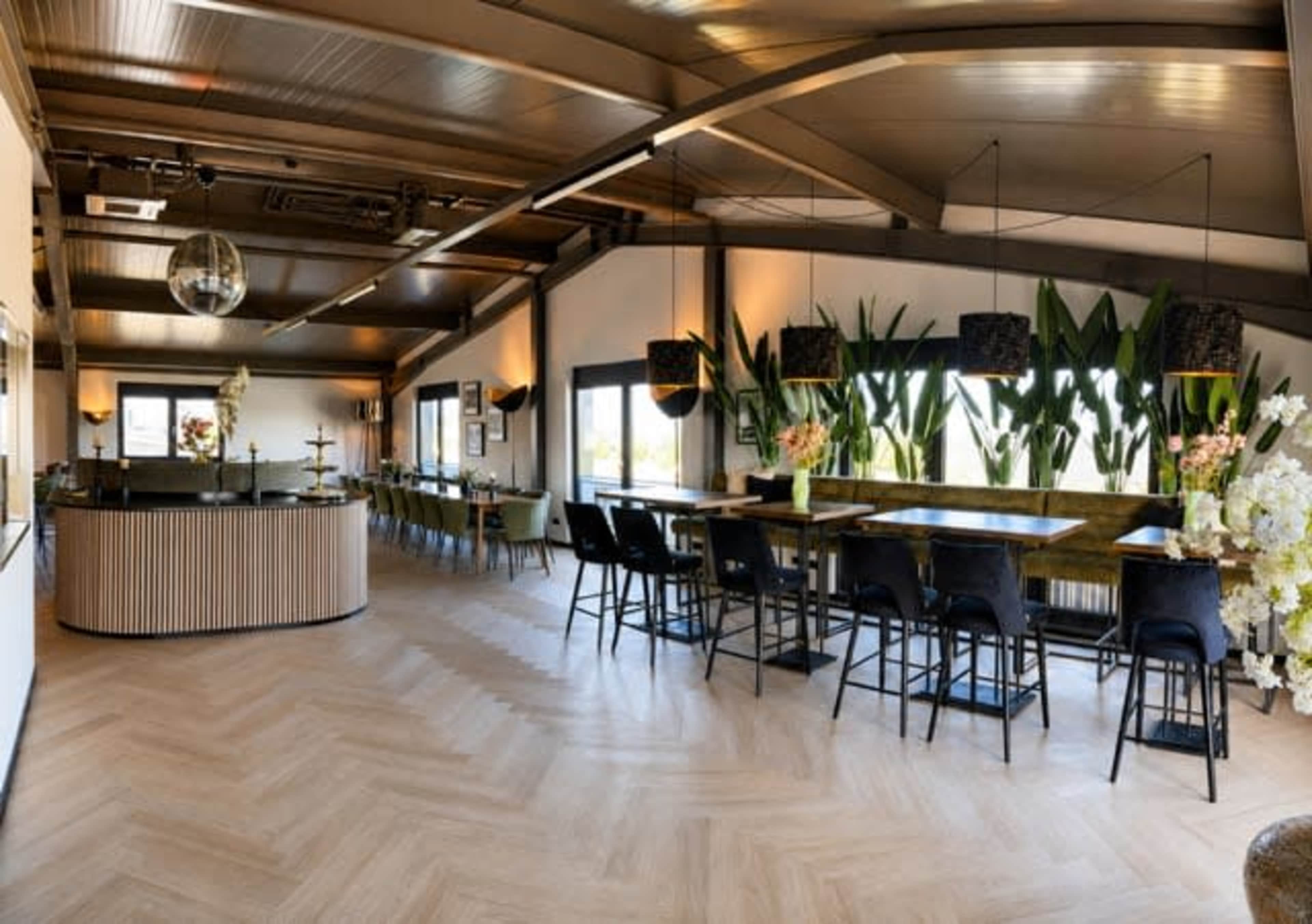 The image shows a modern, spacious dining area featuring a bar counter, tall tables with chairs, and decorative plants throughout.