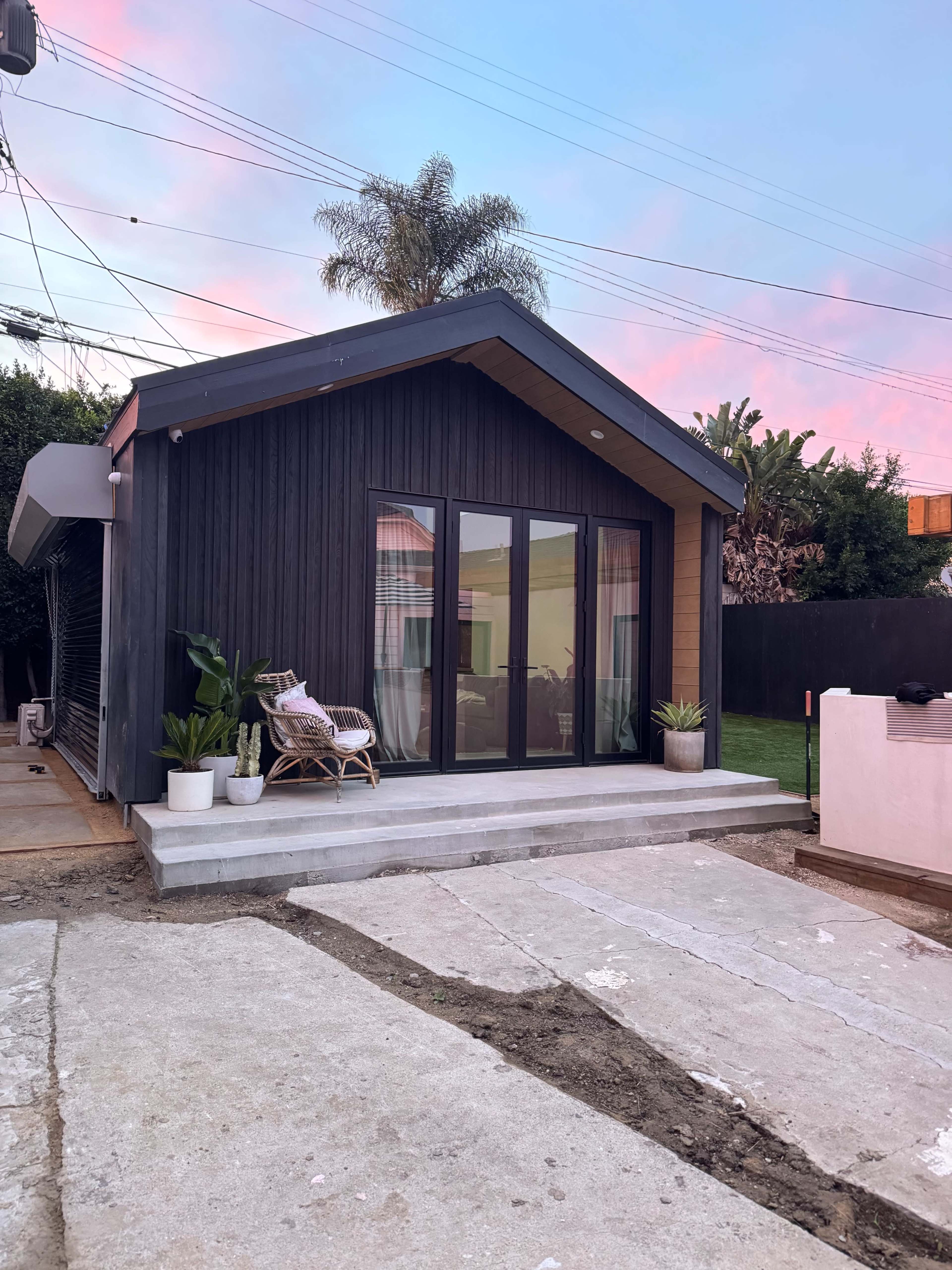 A small, modern house with large glass doors is set against a colorful sunset sky, surrounded by green plants and a clear pathway.