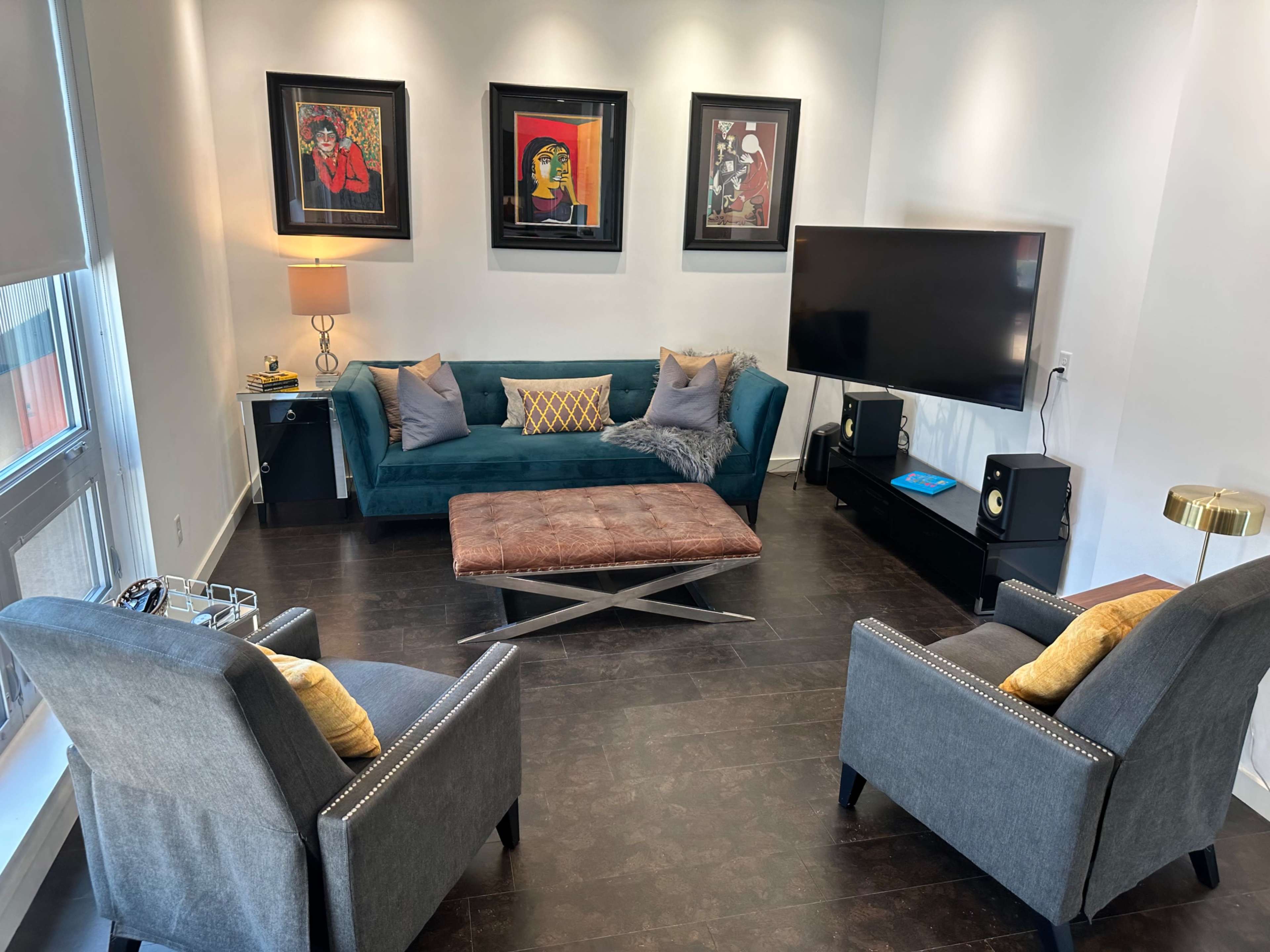 The living room features a teal sofa, two gray armchairs, a coffee table in the center, and framed artwork on the walls, along with a television and a lamp.