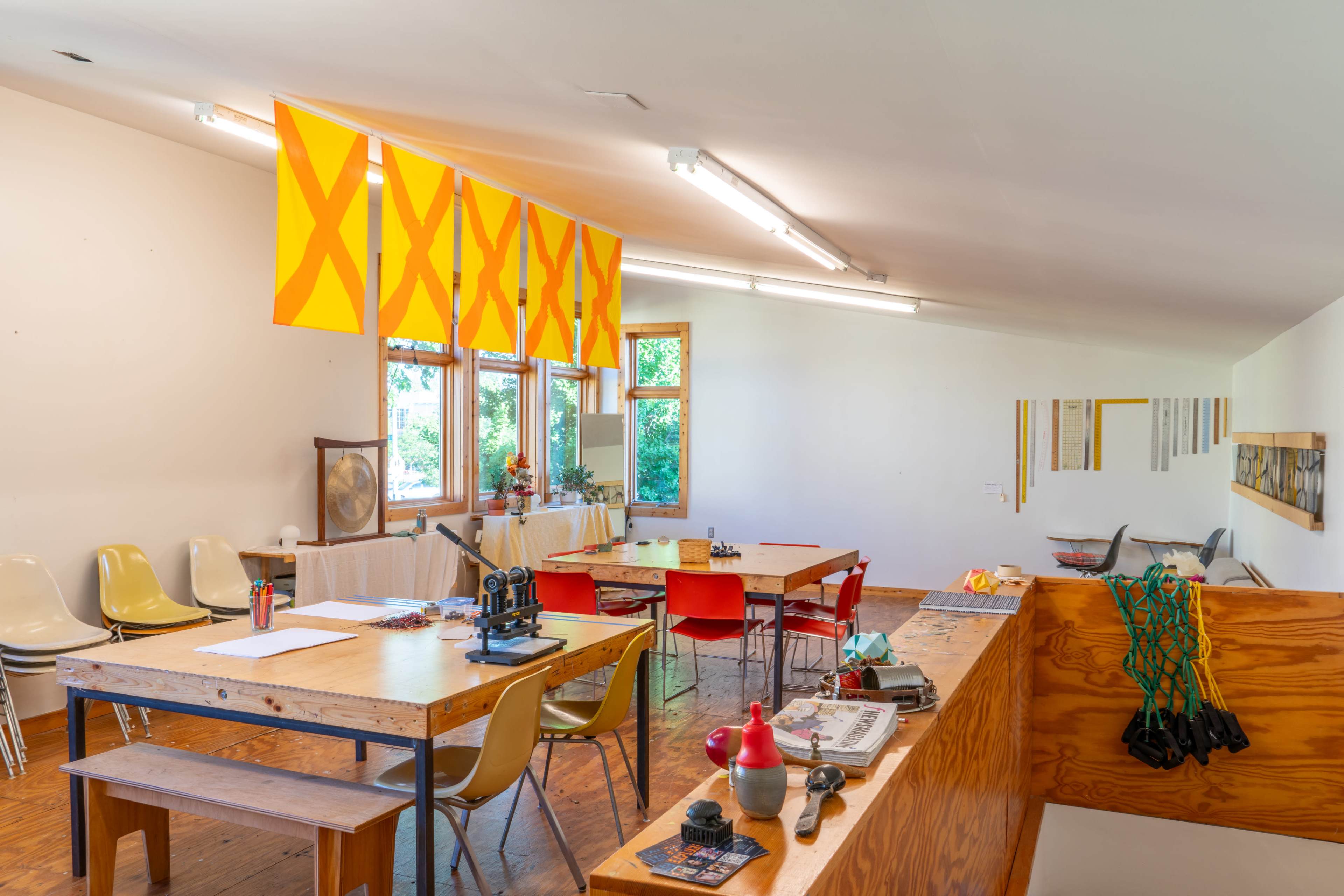 A bright, spacious room with several tables, colorful chairs, large windows, and yellow flags hanging from the ceiling.