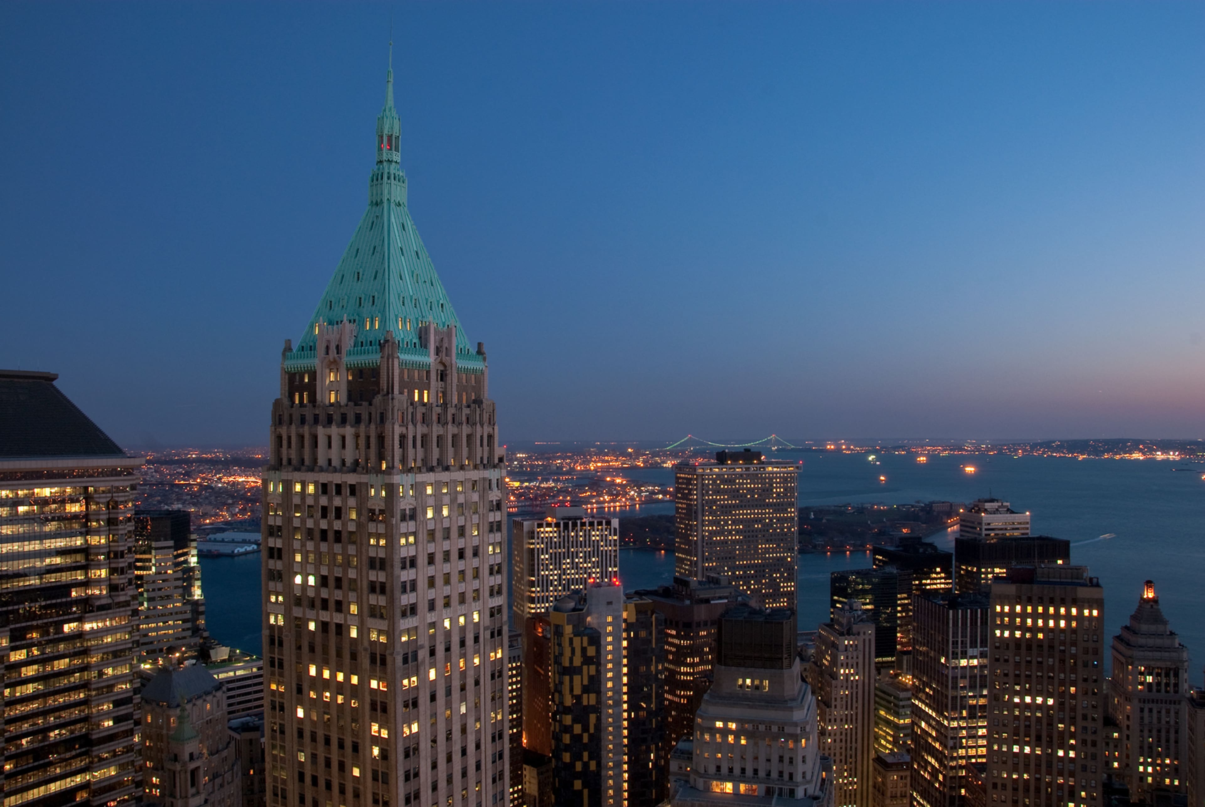The image shows a view of a city skyline at dusk, highlighting a tall building with a green spire and illuminated structures in the background.