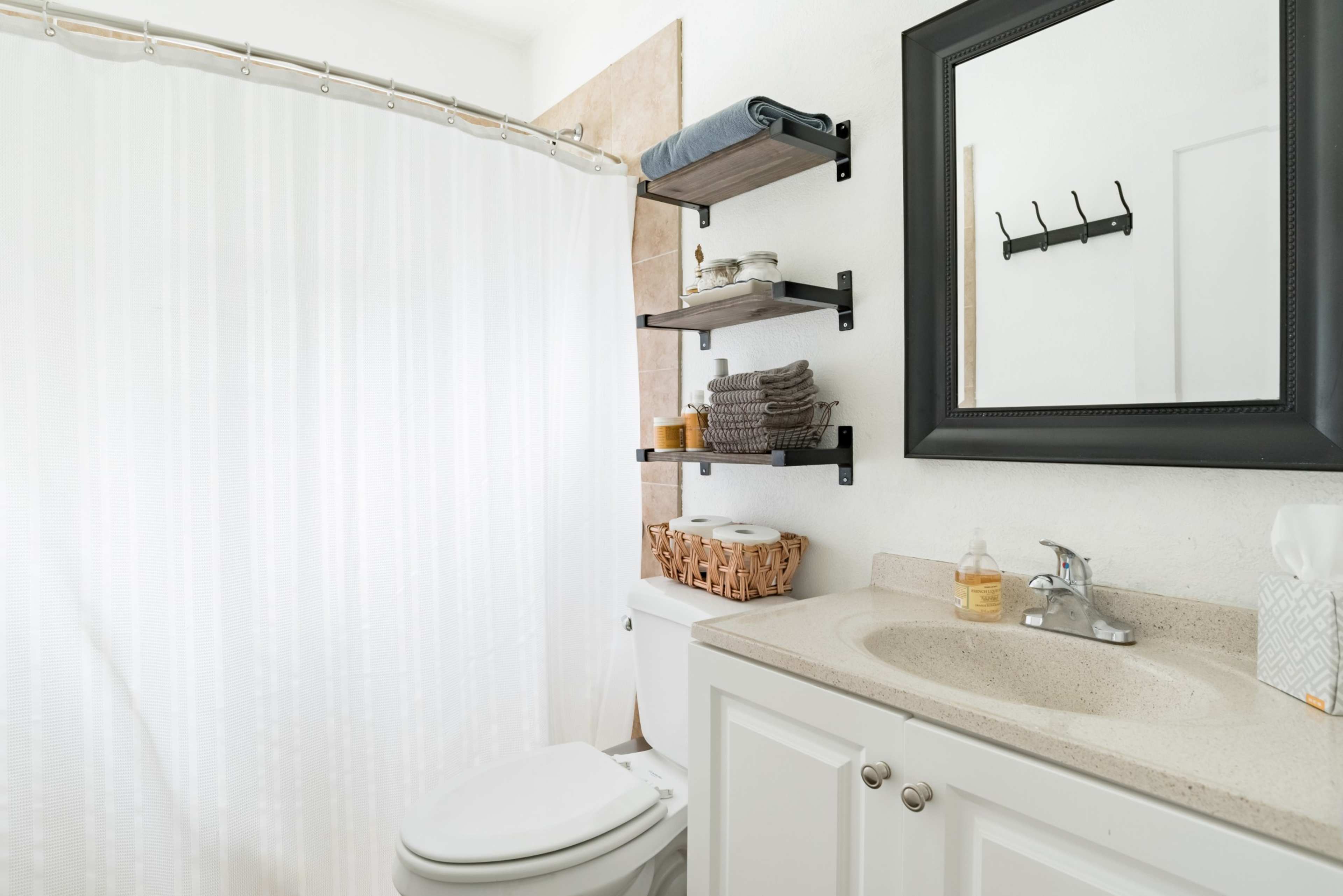 The image shows a clean bathroom with a shower curtain, a sink with a countertop, and shelving holding towels and toiletries.