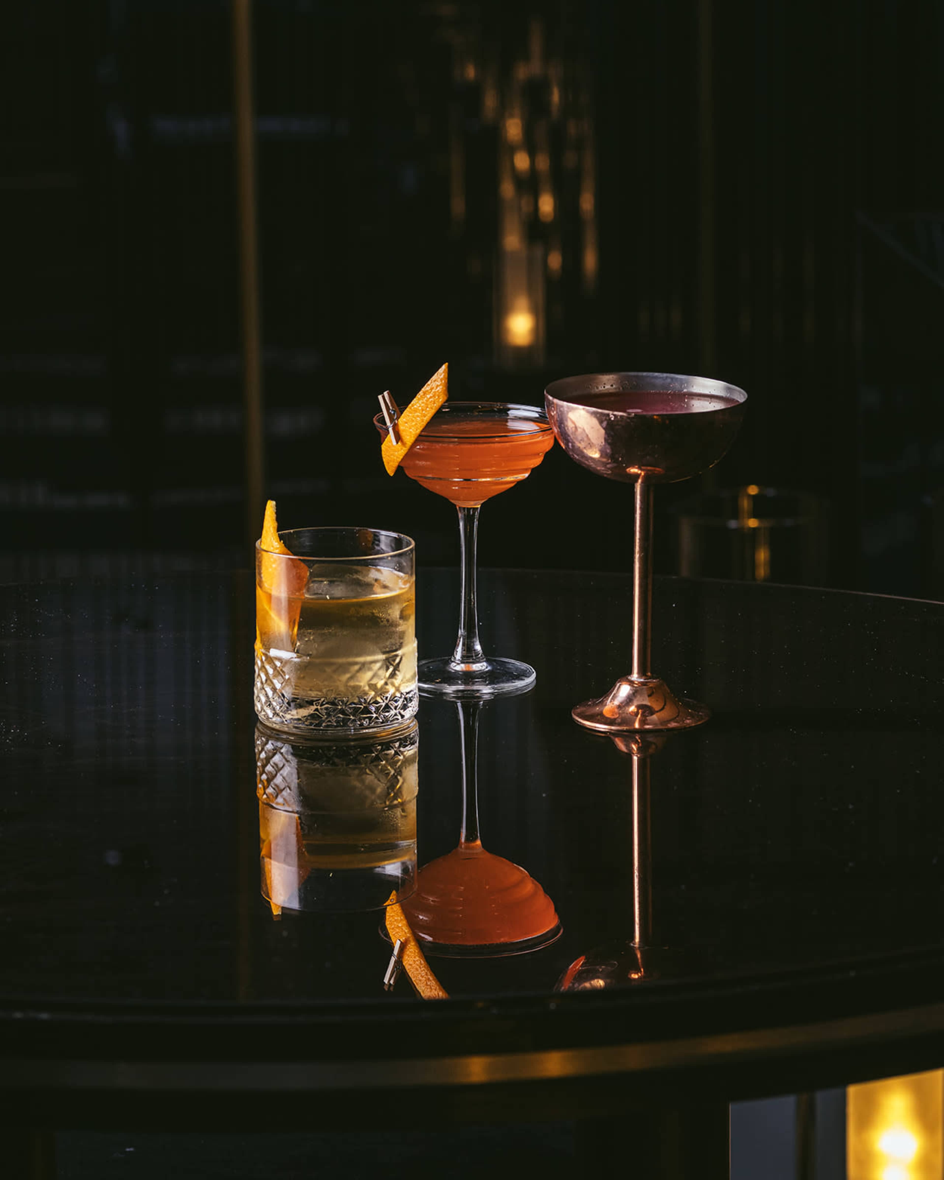 Three cocktails are presented on a dark table, each in distinct glassware, reflecting the surrounding lights.