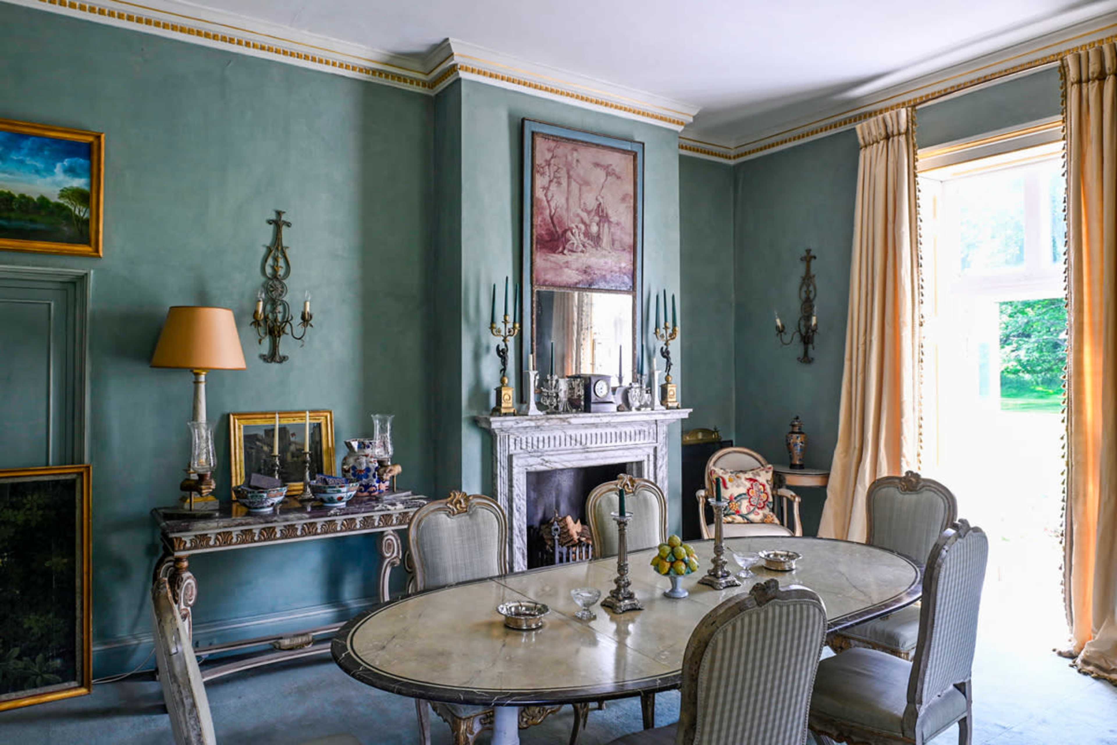 A well-furnished dining room featuring a circular table, antique chairs, a fireplace, and large windows with flowing curtains.