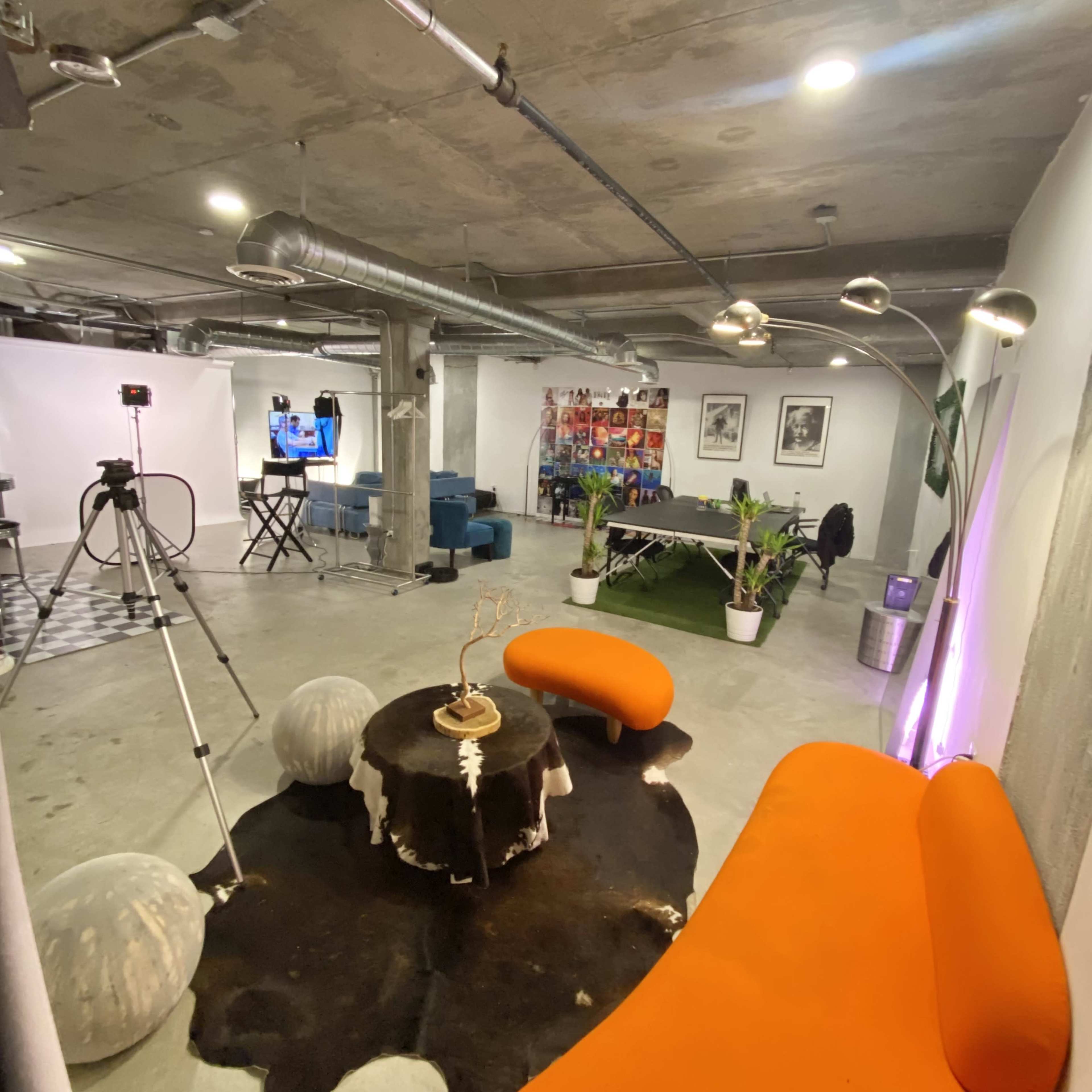 A modern studio space featuring an orange curved couch, a round table on a cowhide rug, a television set, and various seating areas.