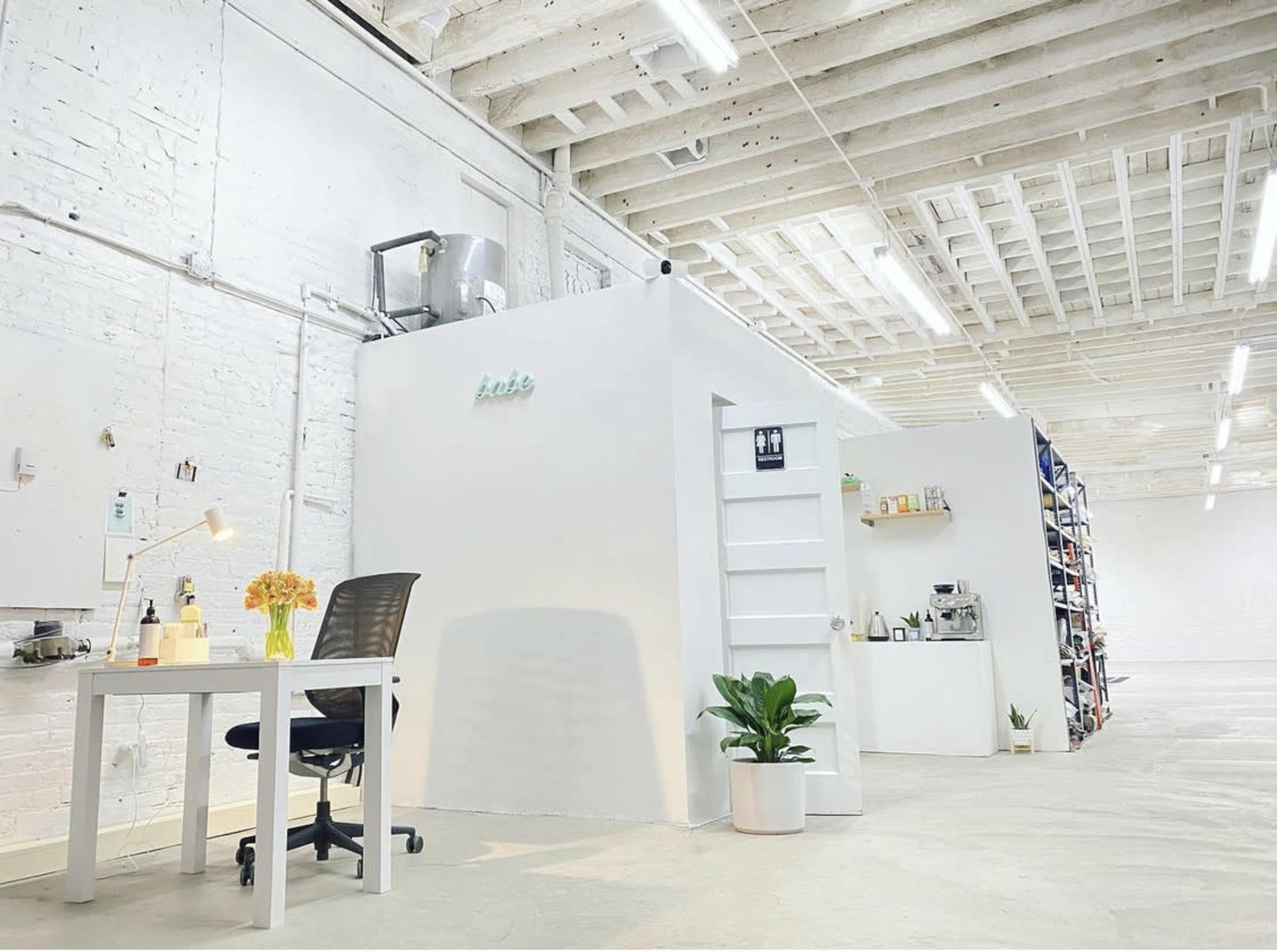 A bright, minimalist workspace featuring a desk with a chair, a potted plant, and shelves stocked with various items against a white wall.