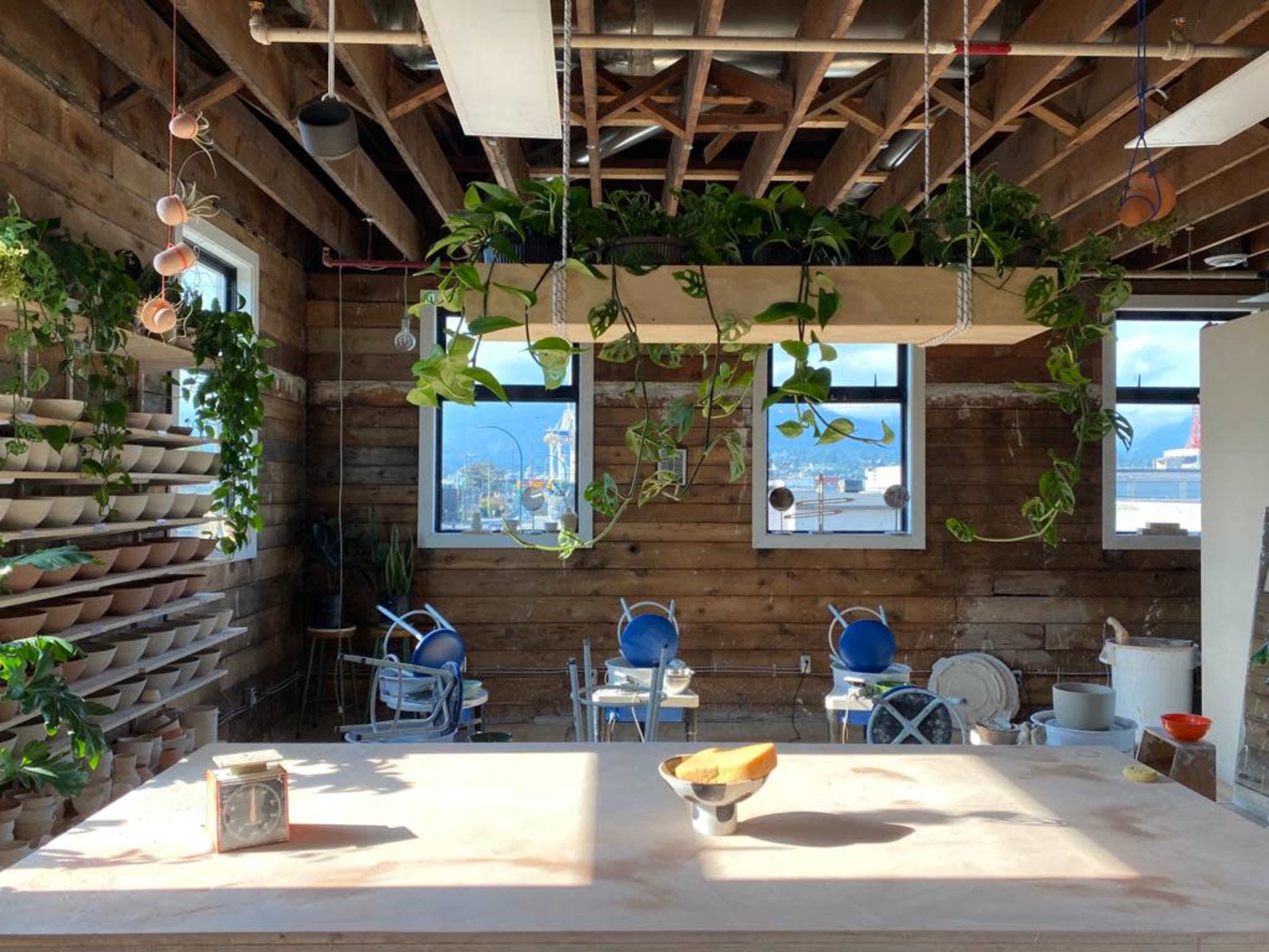 A spacious studio with wooden walls, hanging plants, and a large worktable in the foreground, overlooking a city view through large windows.