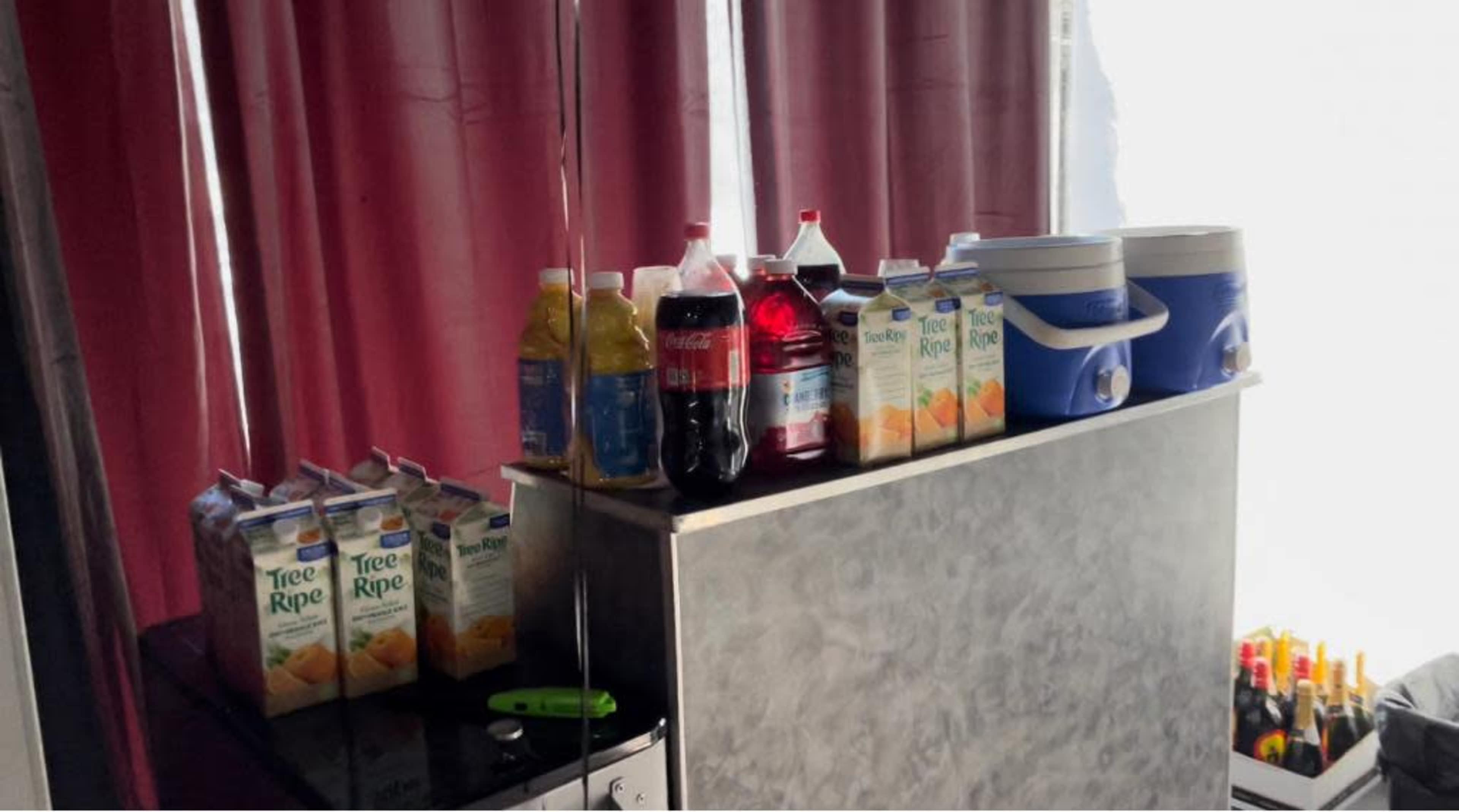 A drink station with various containers and bottles, including juice cartons, soda bottles, and a cooler, set against a backdrop of pink curtains.