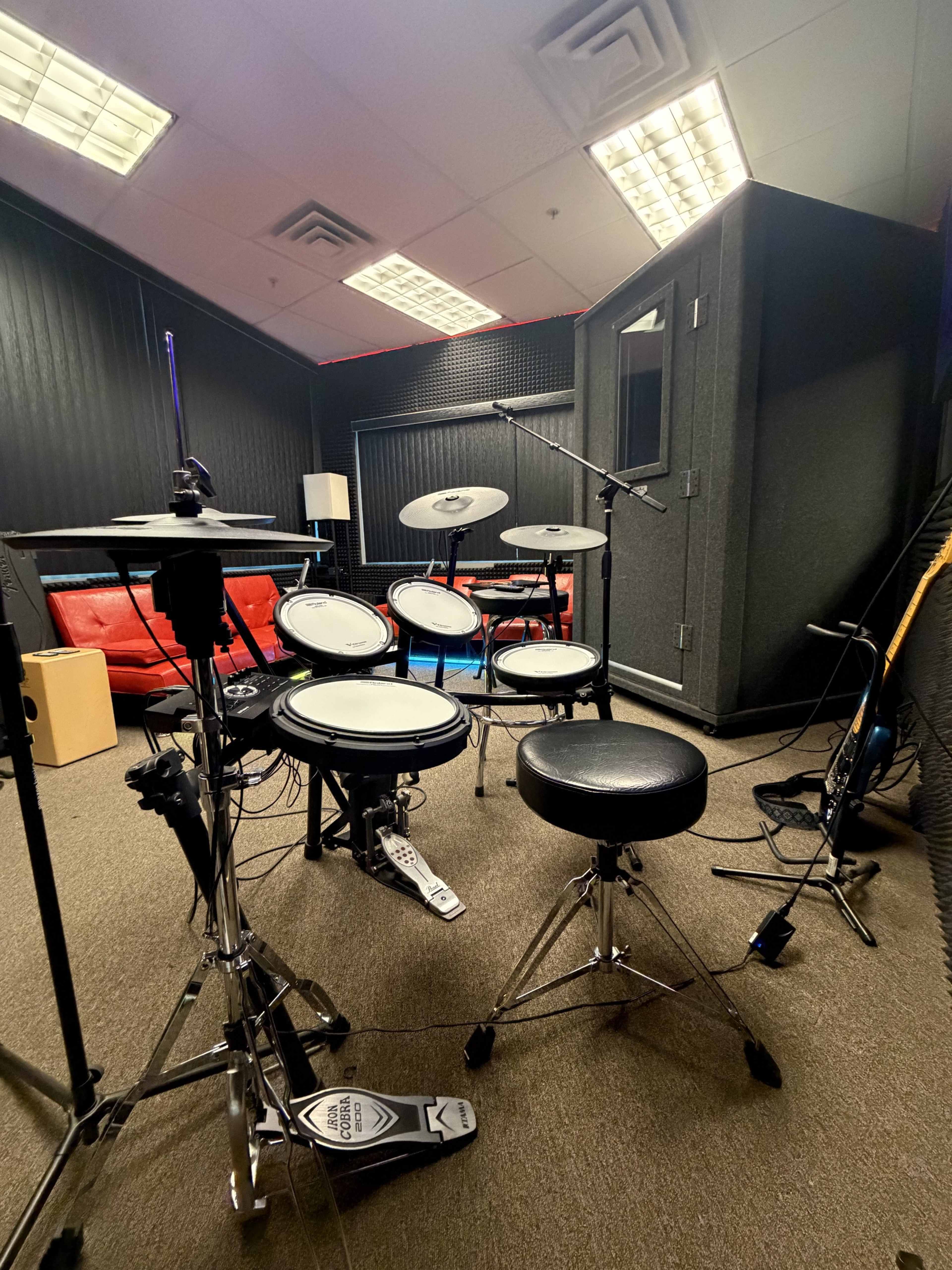 The image shows an electronic drum set positioned in a soundproof studio room with red couches and a wooden box.