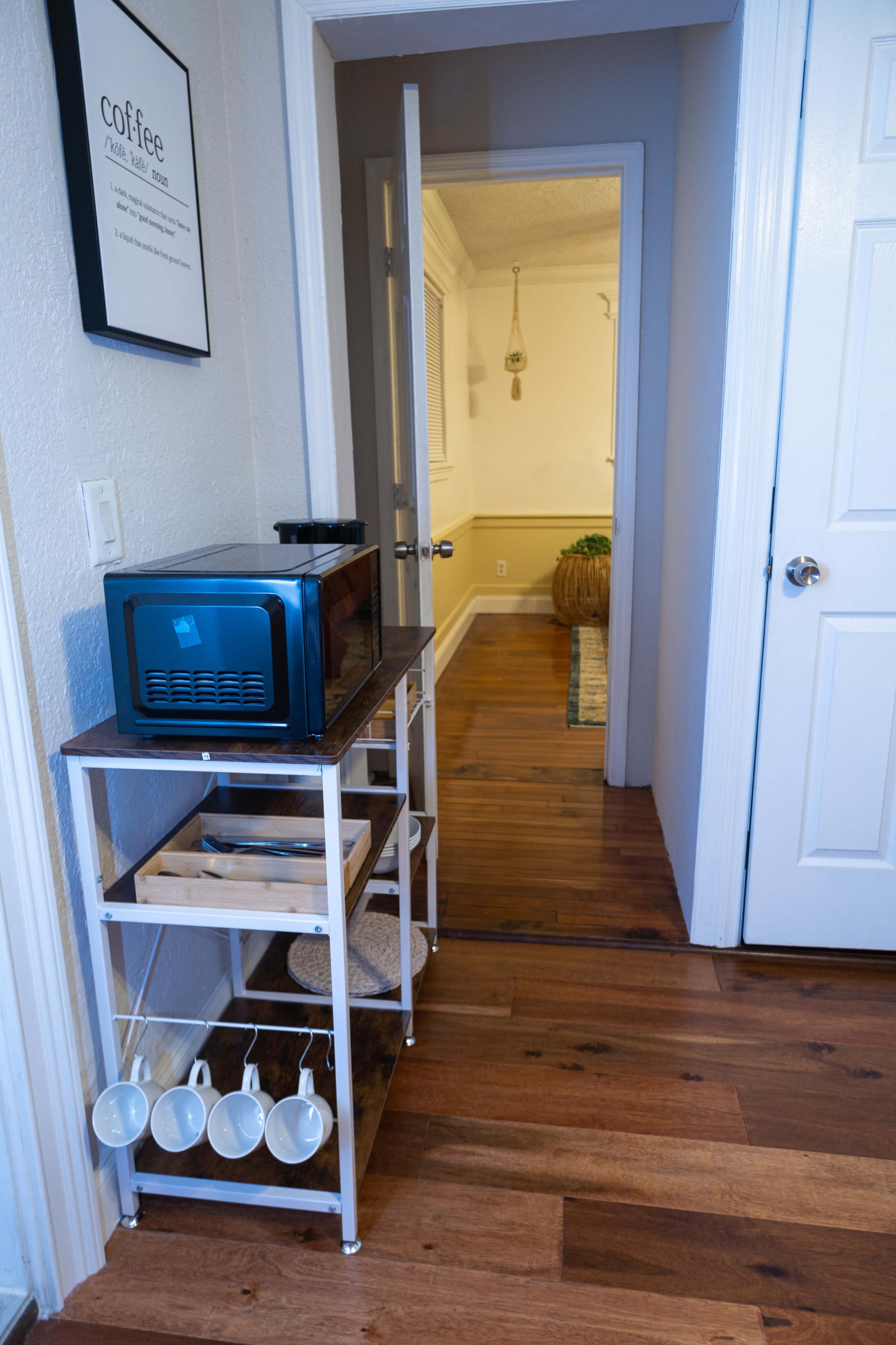 A narrow hallway features a metal kitchen cart with a microwave on top and several hanging coffee mugs, leading to a door that opens into another room.