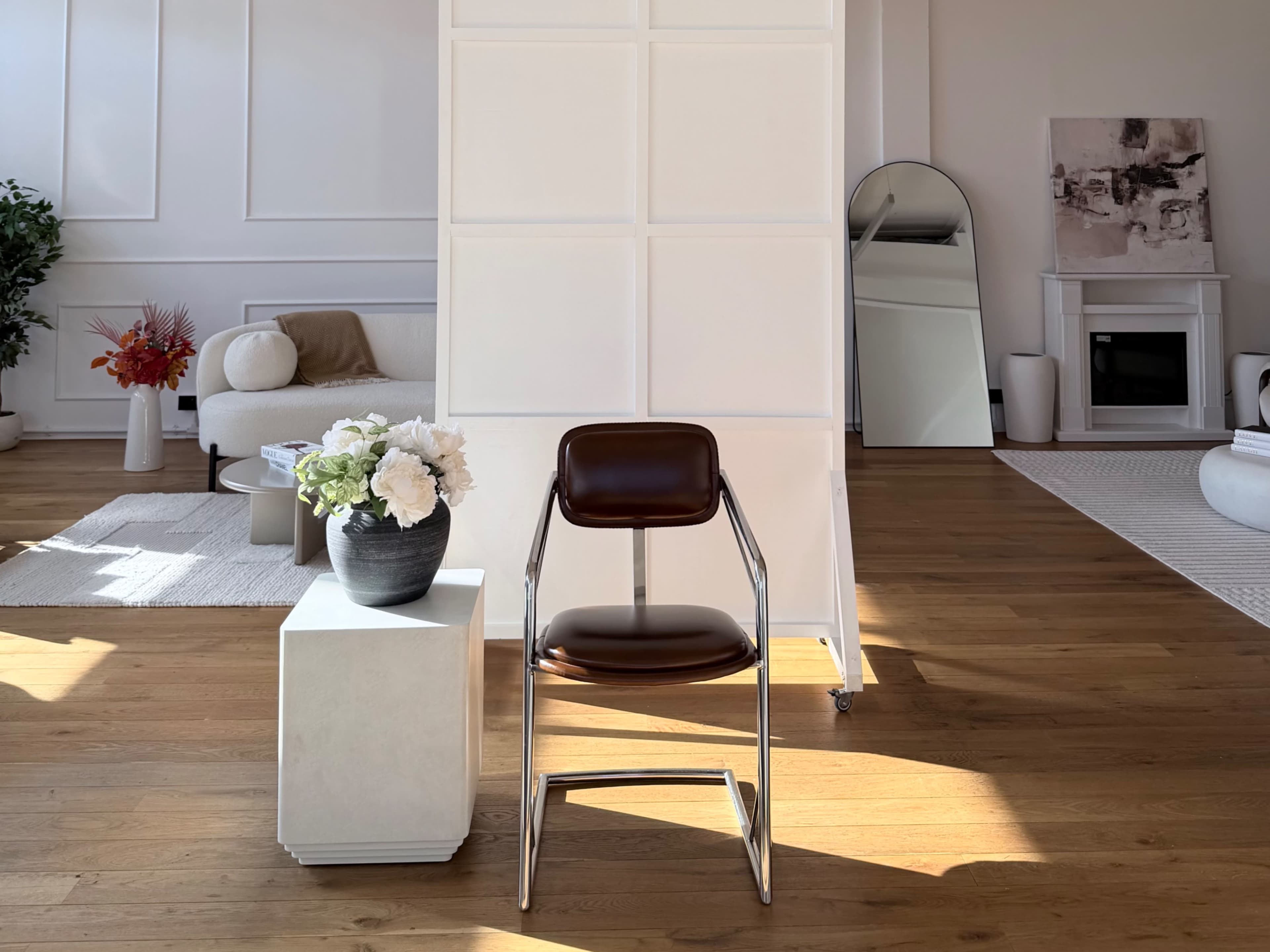 A brown chair is placed next to a white table with a flower vase in a well-lit, modern interior featuring a partition and minimalistic furniture.