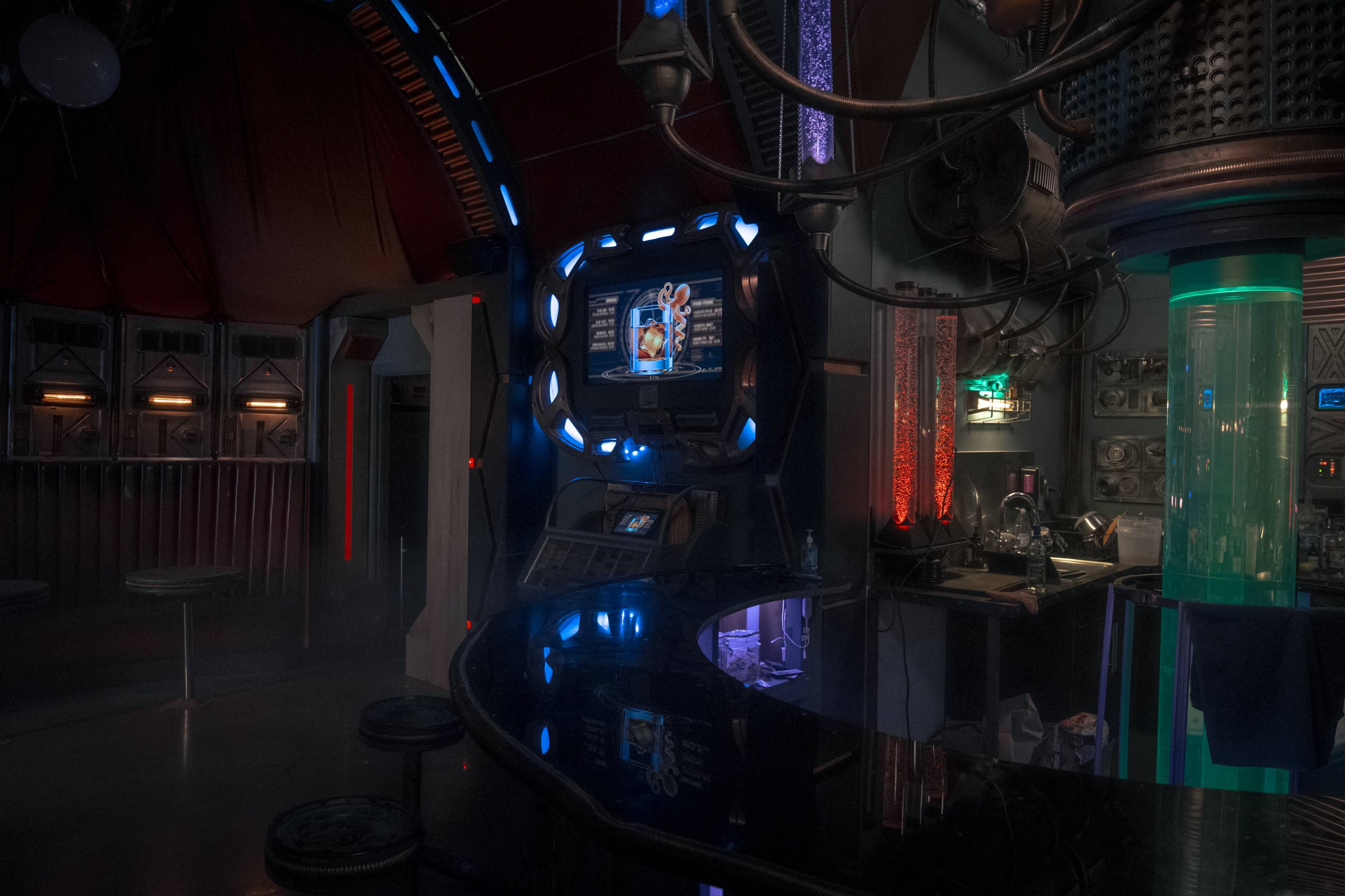 The image shows a futuristic bar interior with vibrant lighting, a curved counter, and a display screen on the wall.