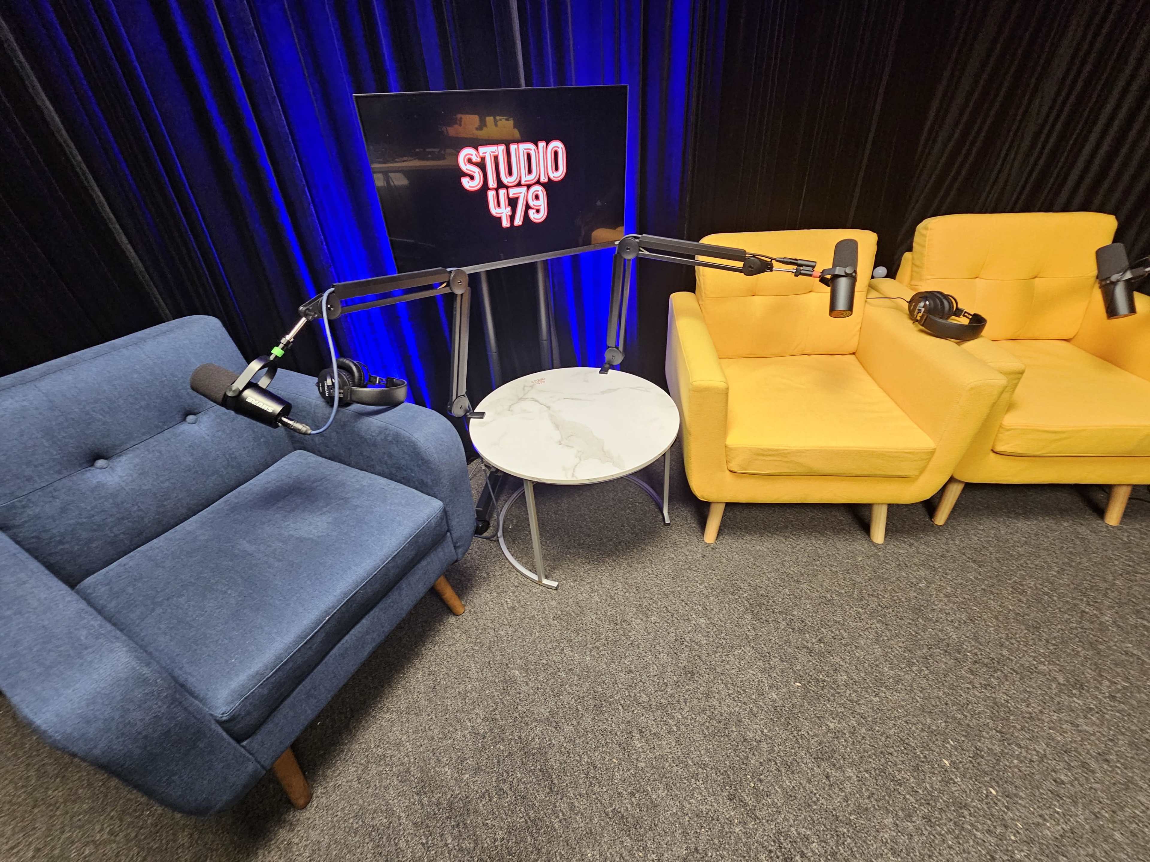 The image shows a small recording studio setup featuring two armchairs, one blue and one yellow, a marble-top table, and a backdrop with the text "STUDIO 479."