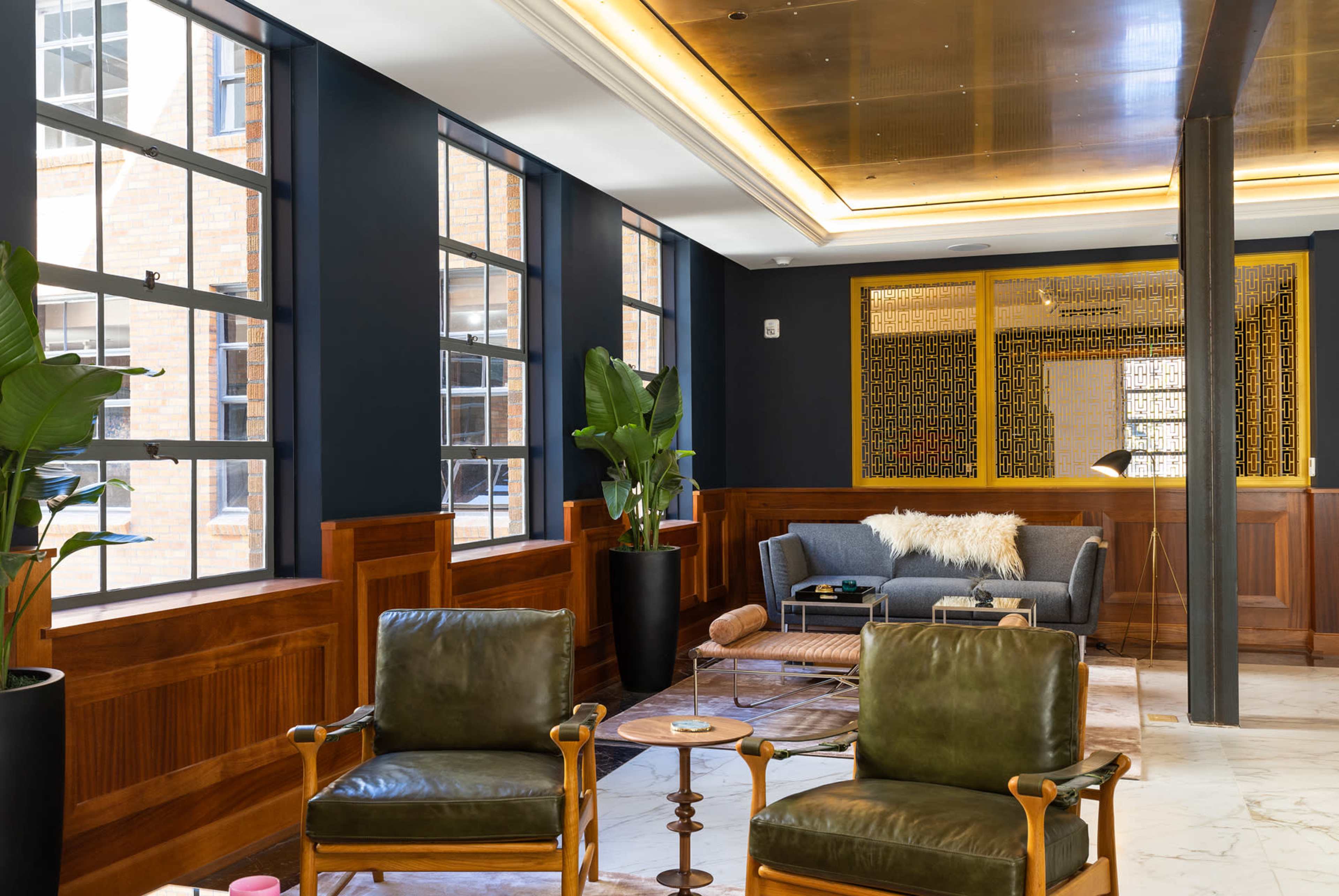 The image shows a modern lounge area with green leather chairs, wooden paneling, large windows, and a sofa in a well-lit space.