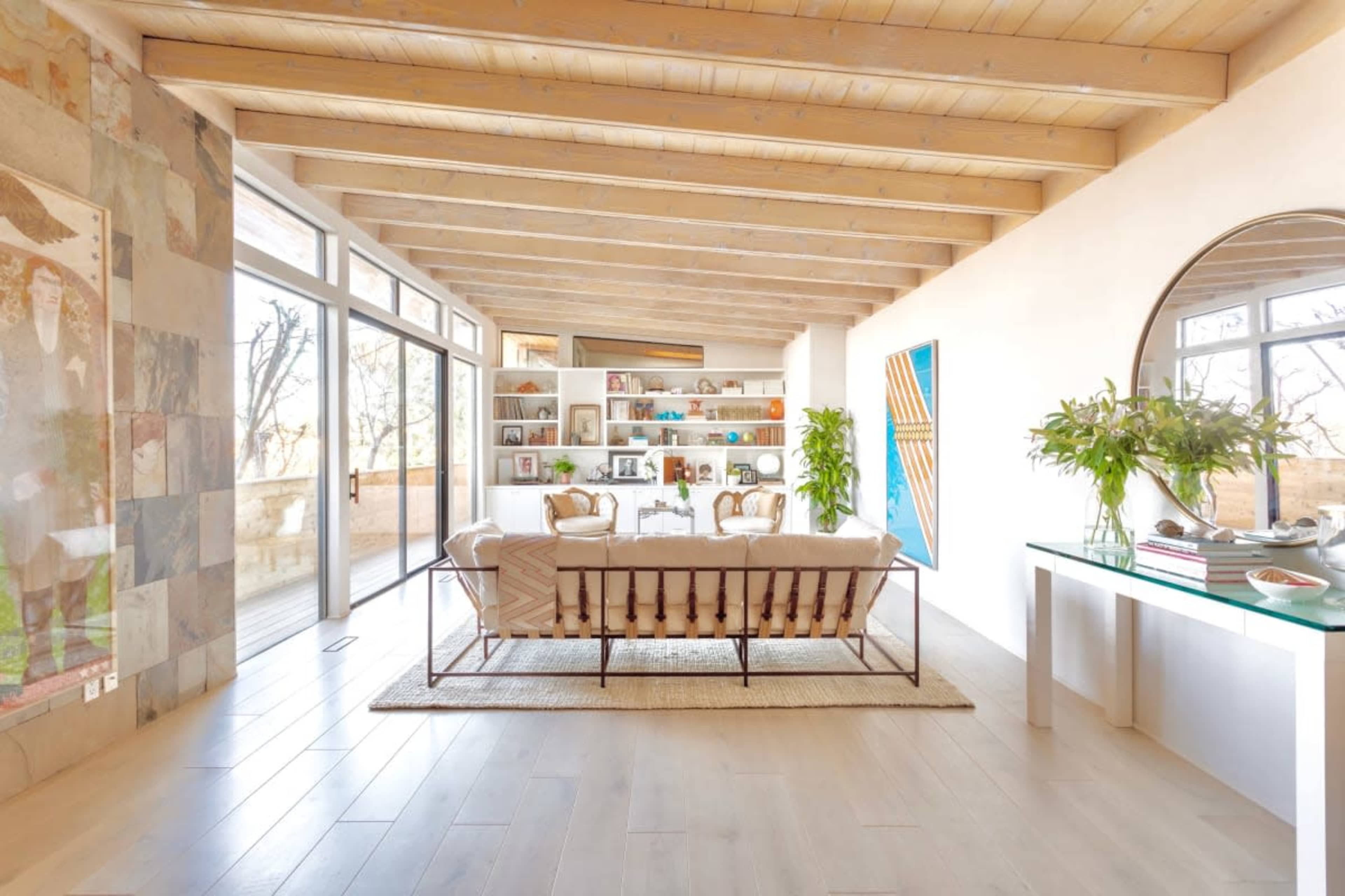 The image shows a modern living room with a beige sofa, a white bookshelf filled with decorative items, and large windows allowing natural light to fill the space.