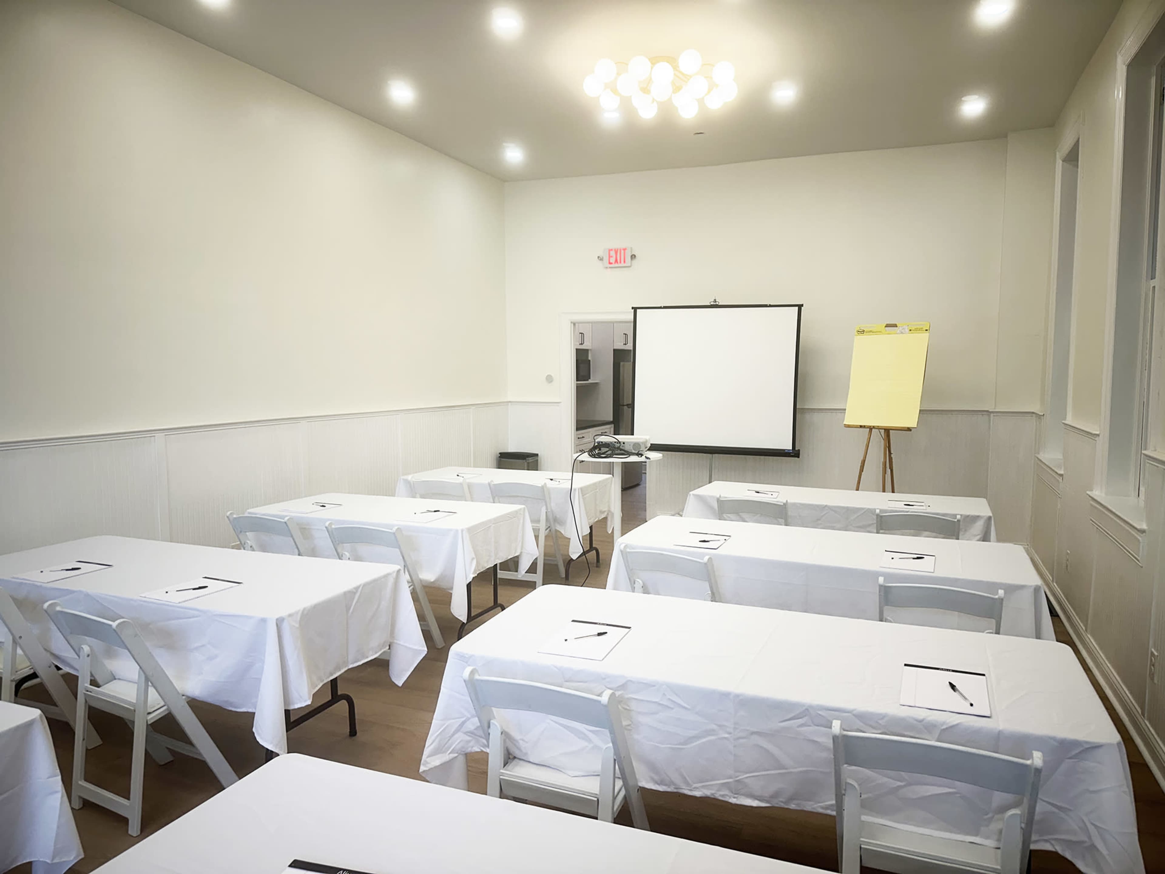 The image shows a bright, empty meeting room with rows of tables covered in white cloths and a projector screen at the front.