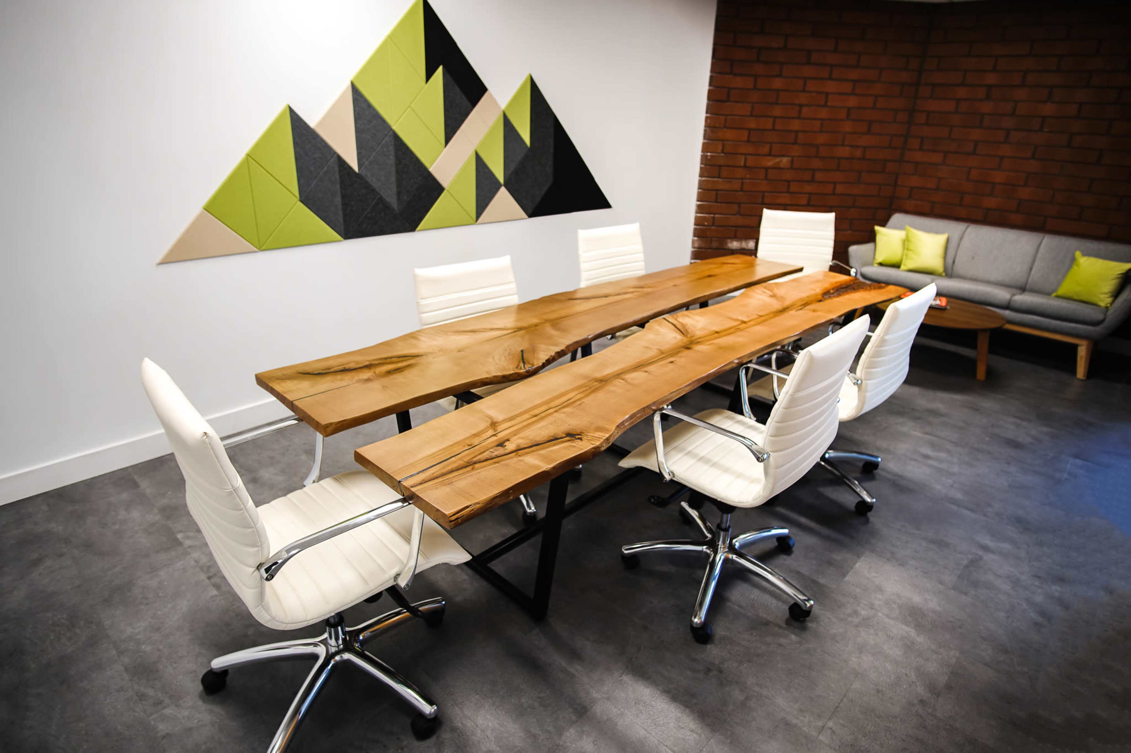 A wooden conference table with a live edge design is surrounded by four white office chairs in a meeting room featuring a geometric wall decoration and a gray couch.