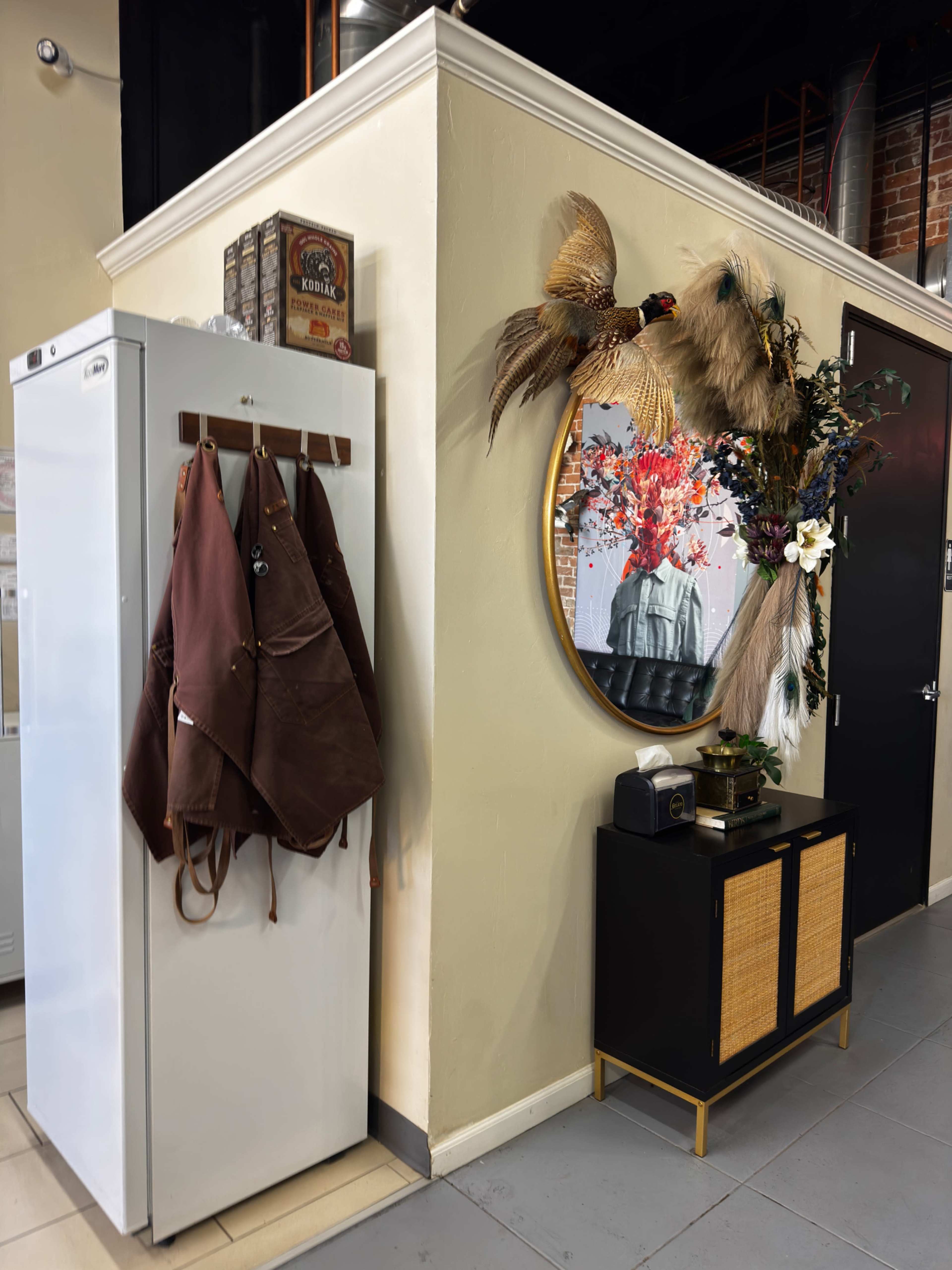 A white refrigerator is situated next to a mirrored wall adorned with a decorative arrangement of dried plants and flowers, with a black and rattan cabinet below it.