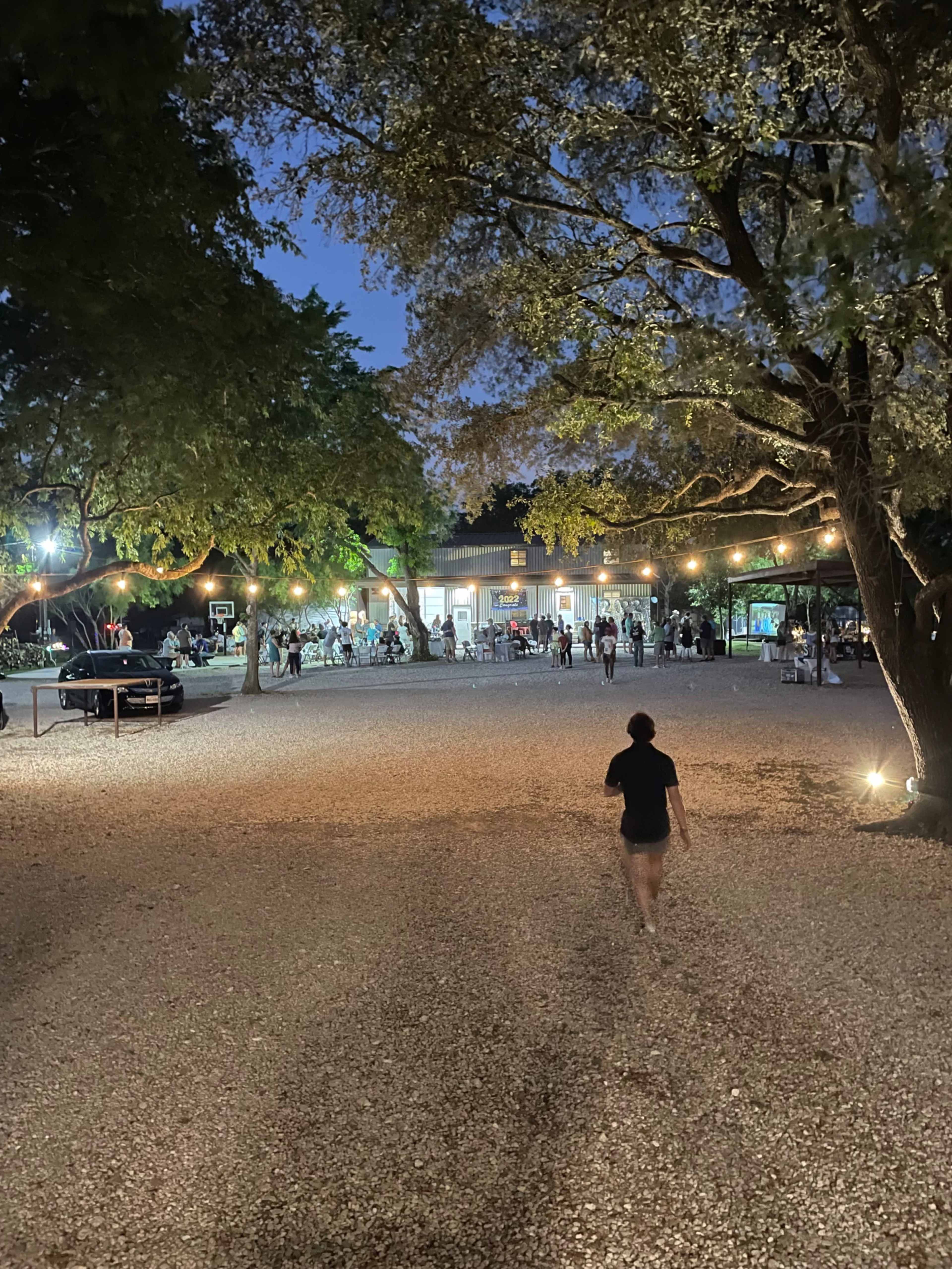 A person walks along a gravel path toward a well-lit gathering area surrounded by trees and decorated with string lights at dusk.