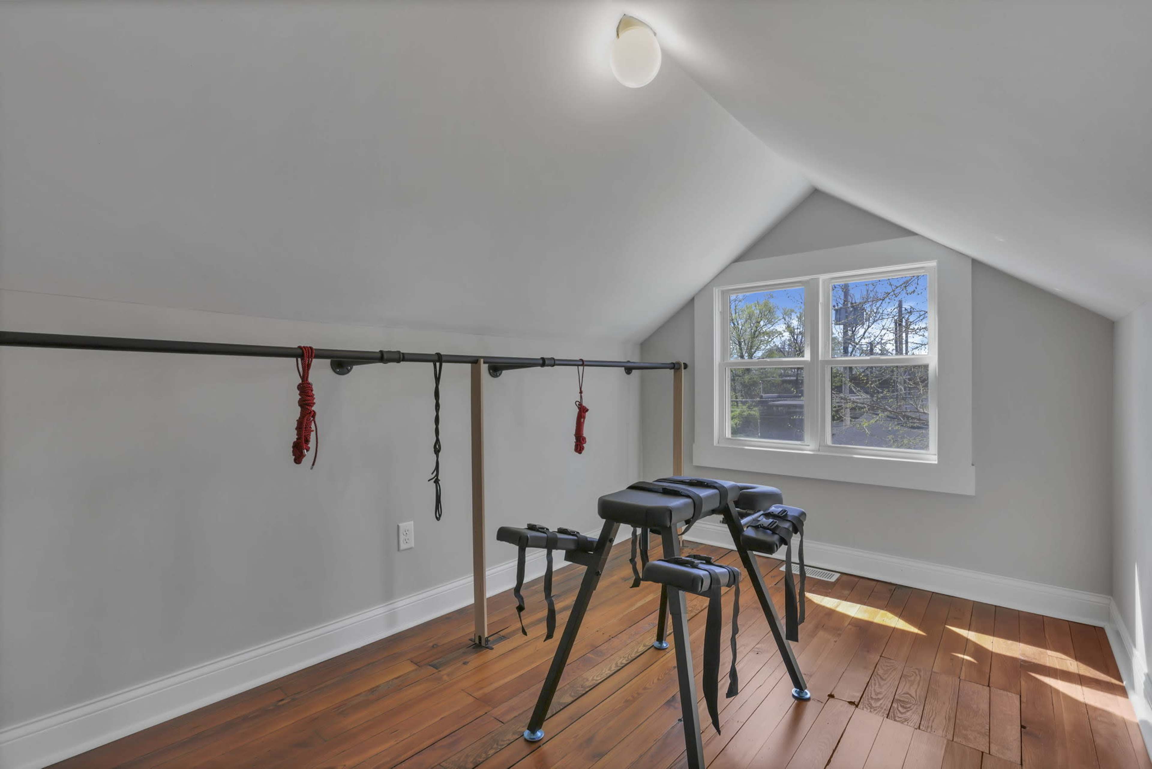 The image shows a small attic room with wooden floors, featuring a set of exercise bars and a workout bench under a slanted ceiling with a window allowing natural light.