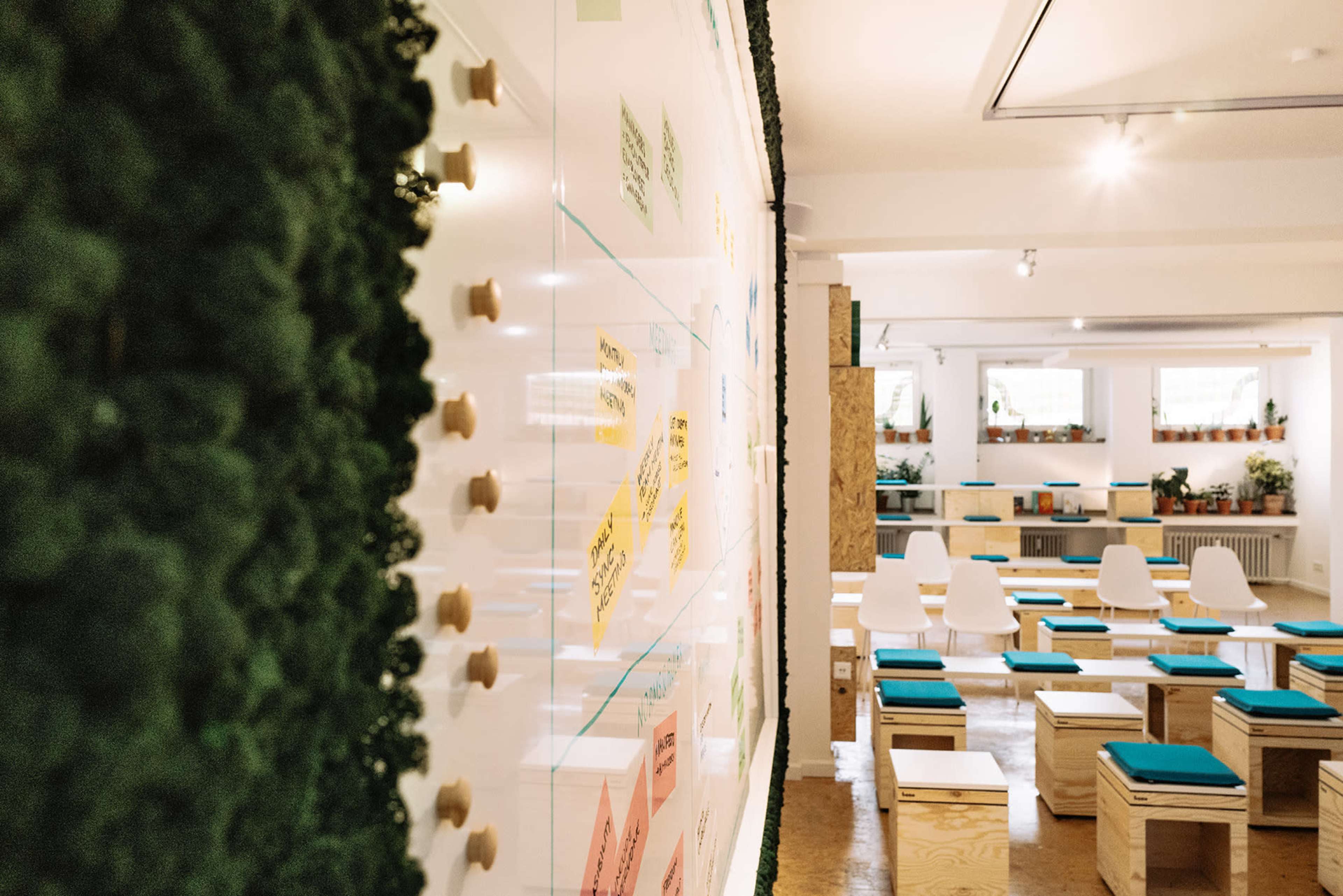 A modern workspace with a whiteboard featuring colorful sticky notes, surrounded by wooden seating arrangements and greenery.