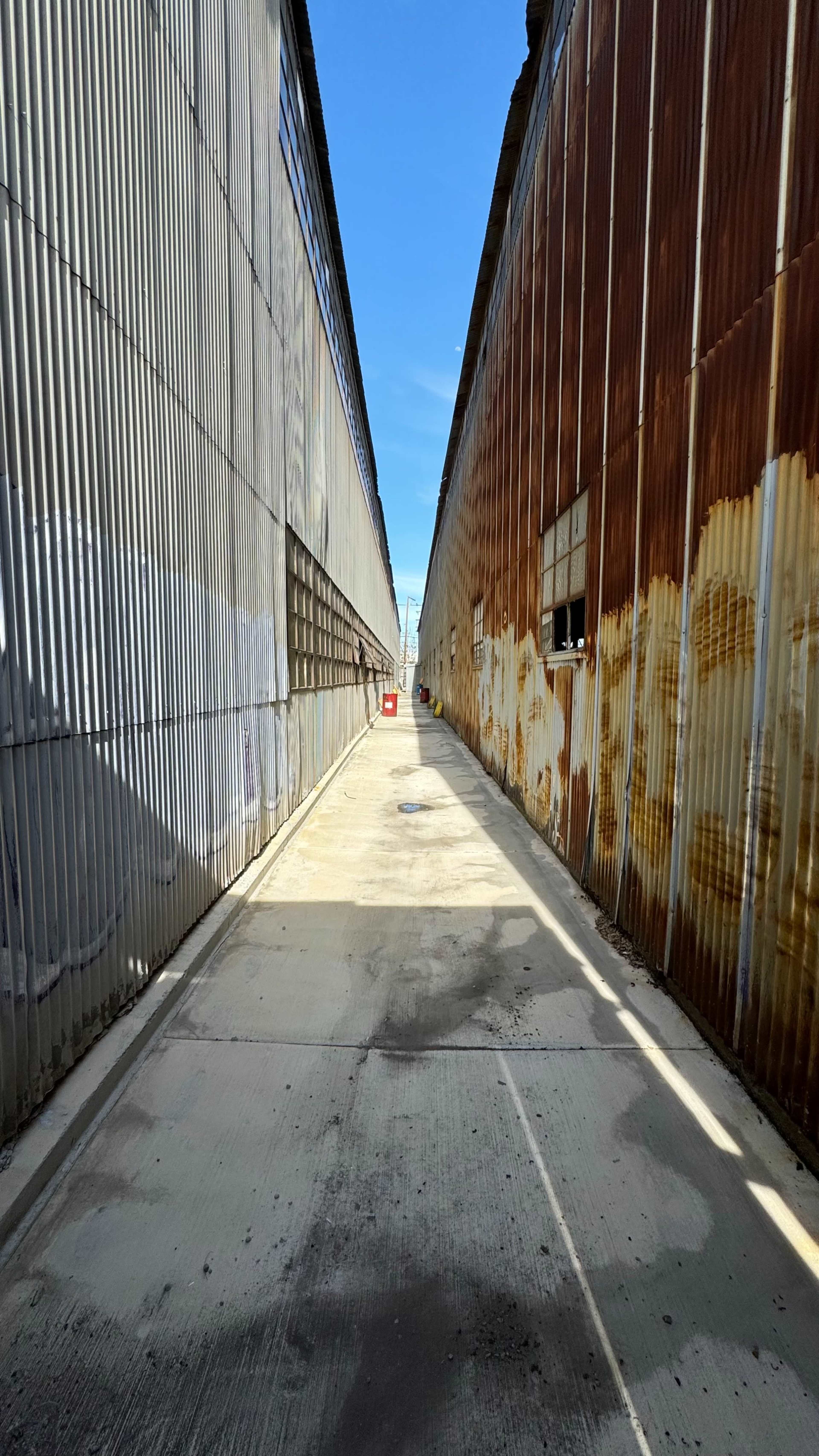 A narrow alleyway flanked by two metal walls, one smooth and silver, the other rusted and orange, under a clear blue sky.